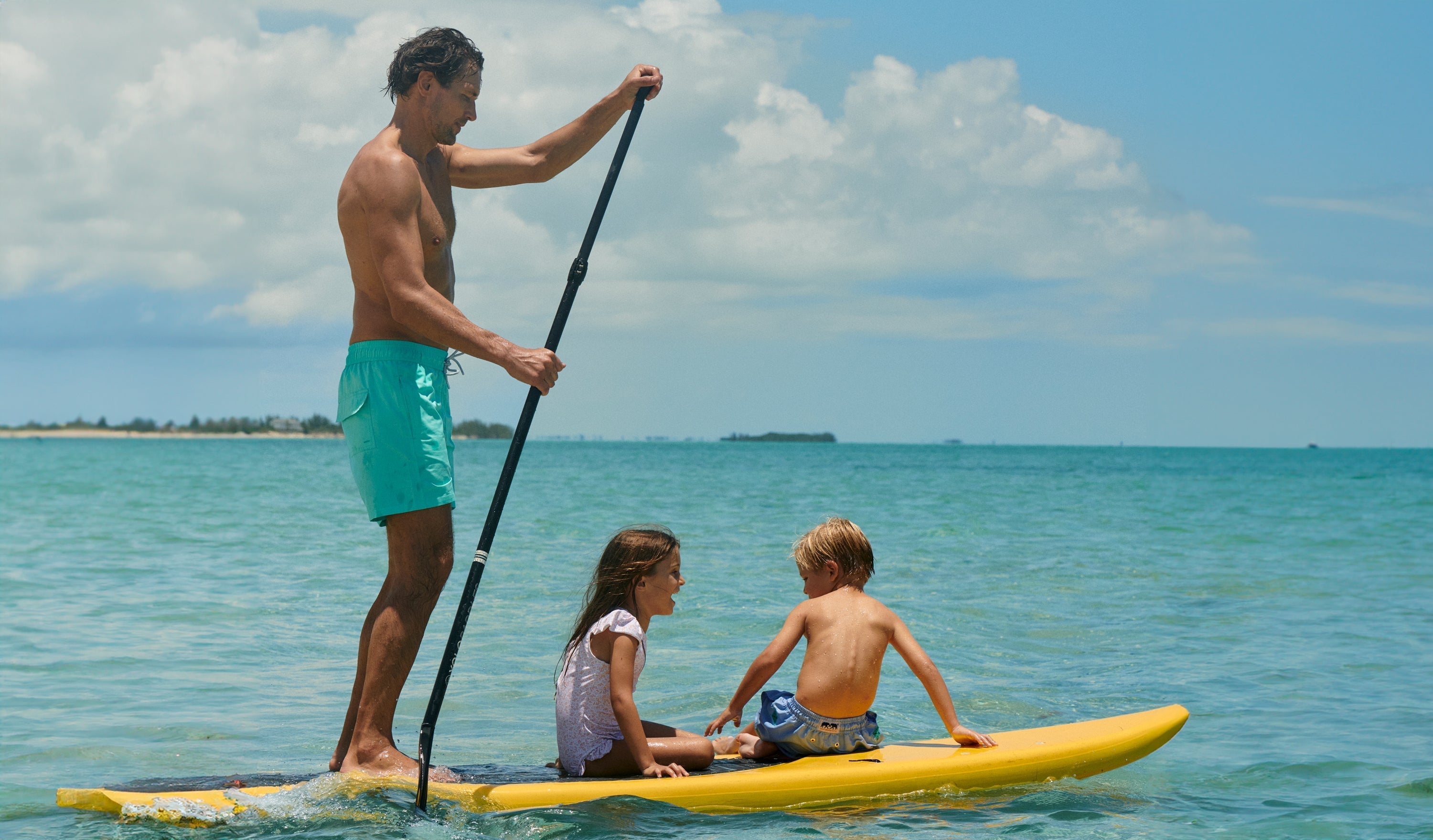 Man wears swim shorts on a paddle-board with 2 children.