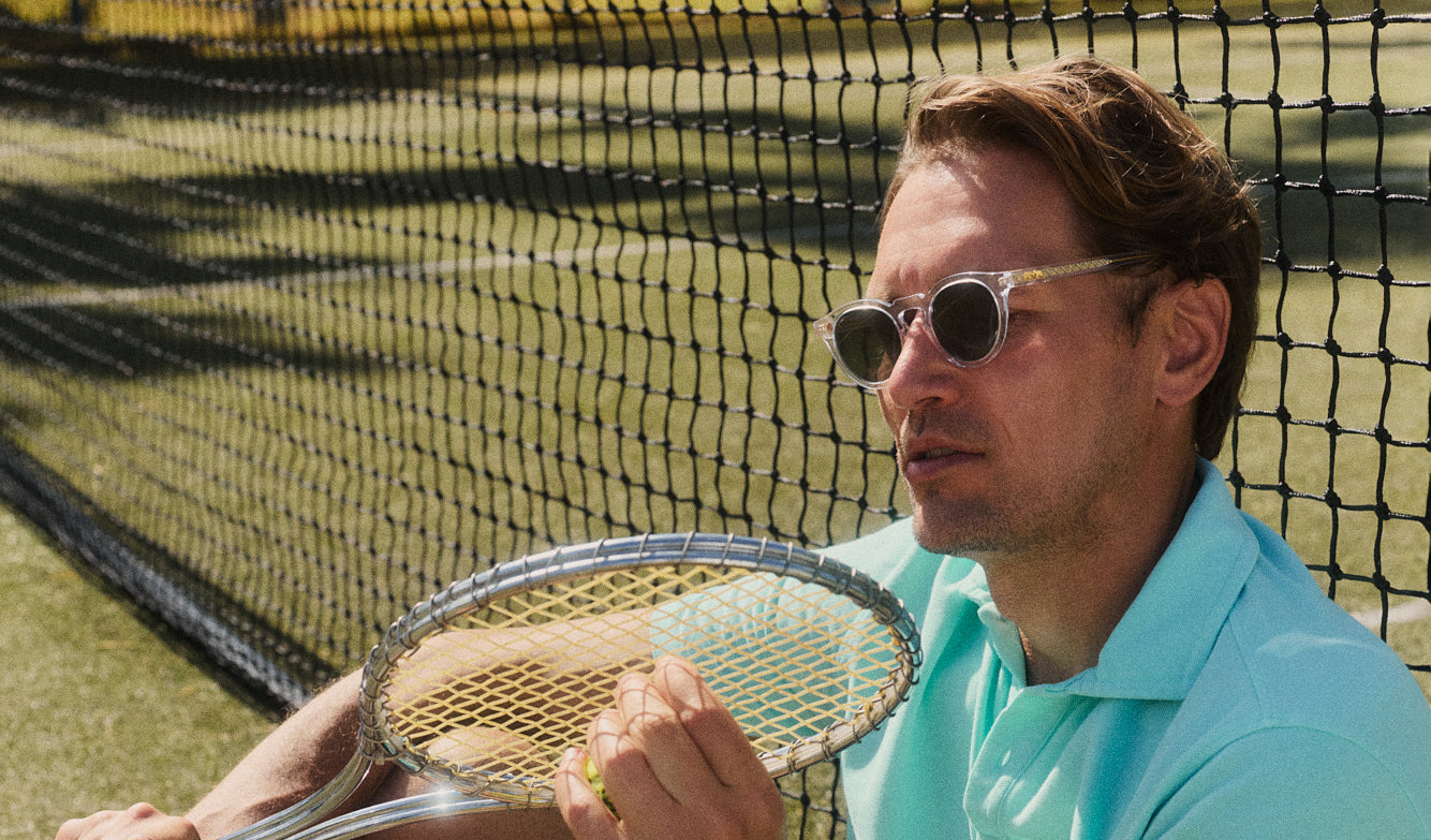 Man wears clear sunglasses with brown lenses while leaning on a tennis net holding a tennis racket.