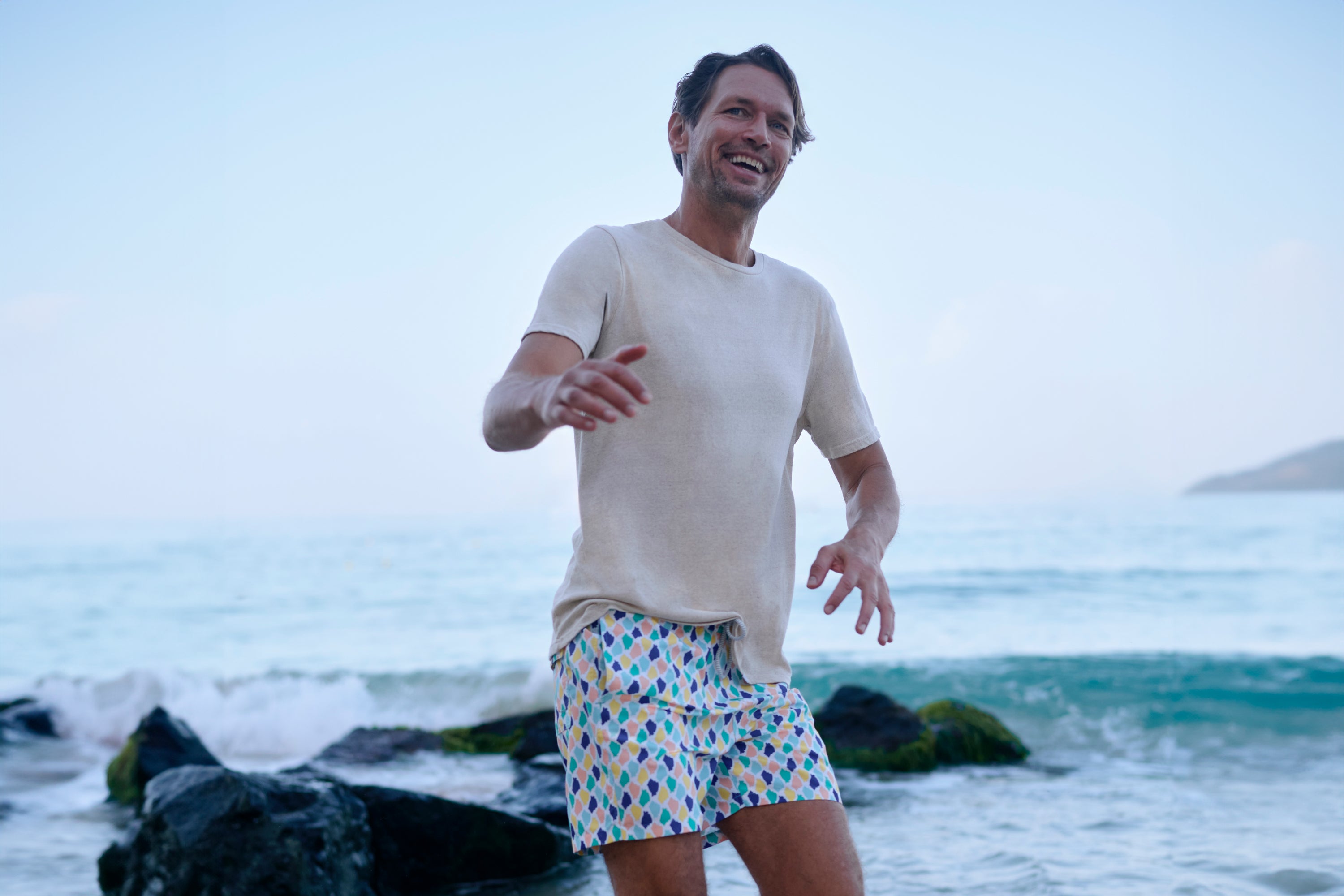 Man standing on a beach with ocean and sky in the background wearing multicoloured swim trunks