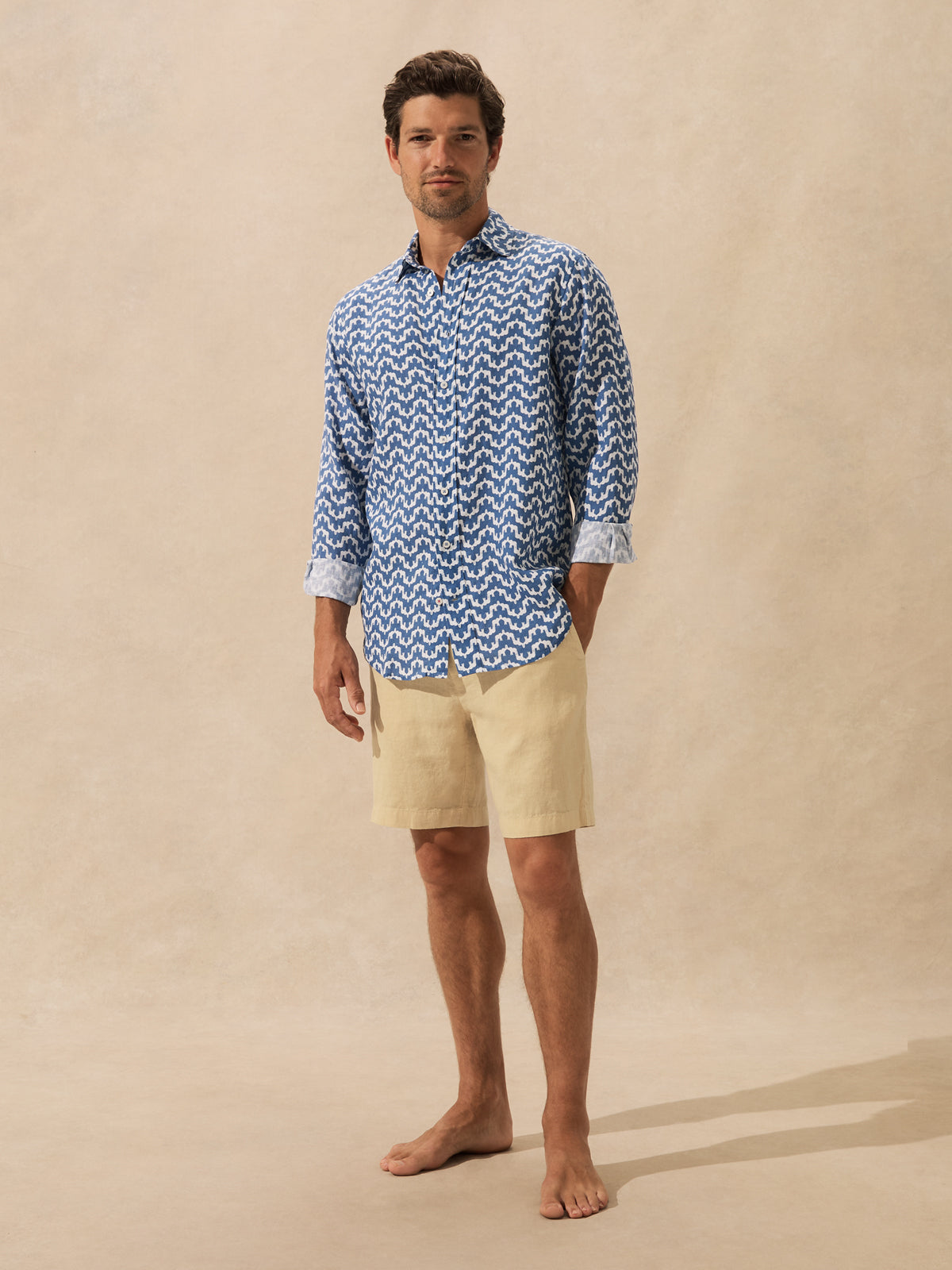 A man stands barefoot against a warm beige studio backdrop, wearing a long-sleeve button-up shirt with a blue and white elephant pattern and light beige shorts.