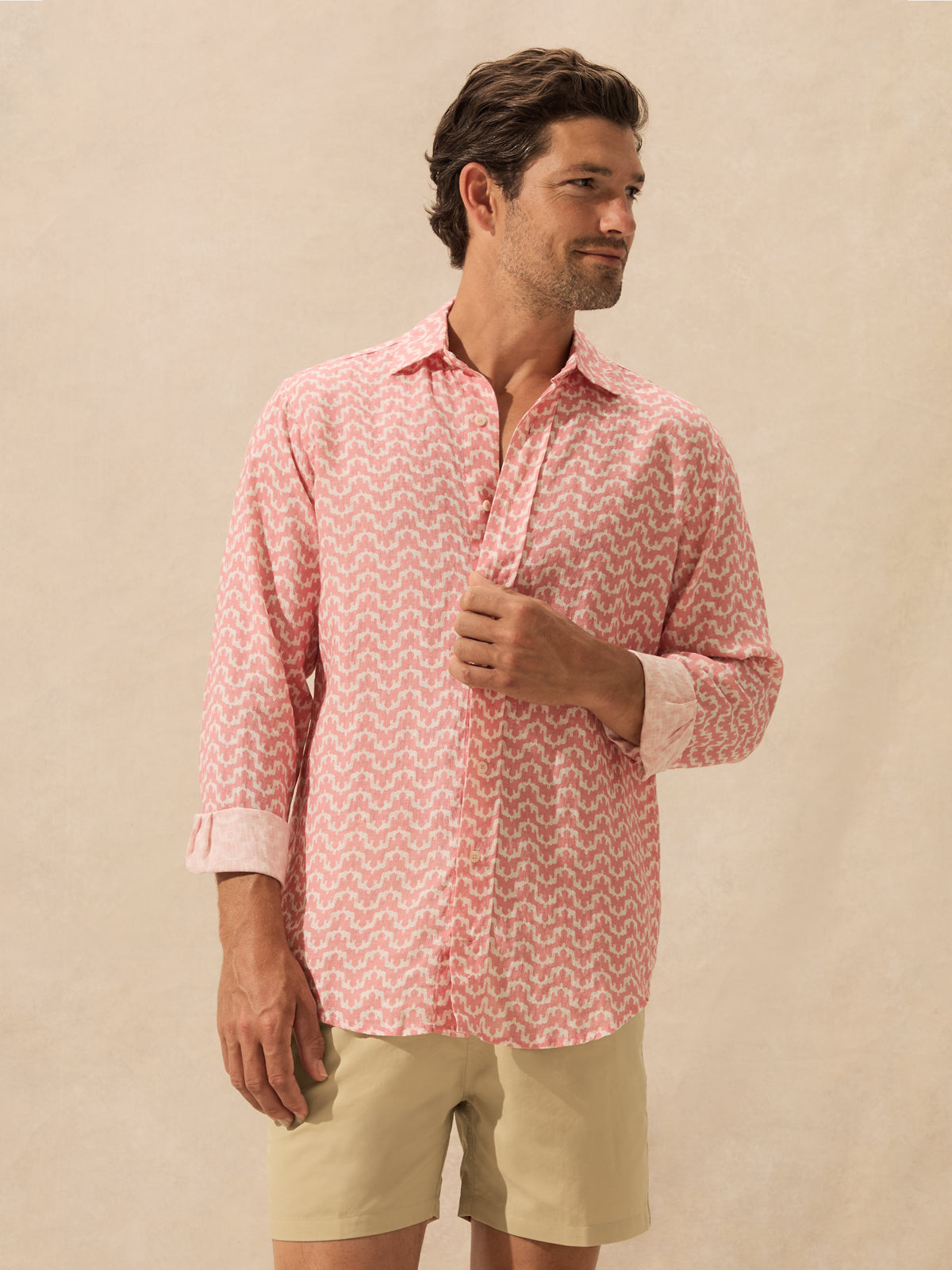 A man wearing a pink and white patterned long-sleeve button-down shirt standing against a textured beige background.