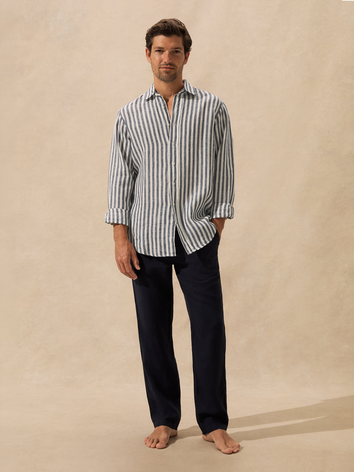 A man stands barefoot against a warm beige studio backdrop, wearing a long-sleeve button-front shirt with narrow vertical blue and white stripes and dark navy pants.