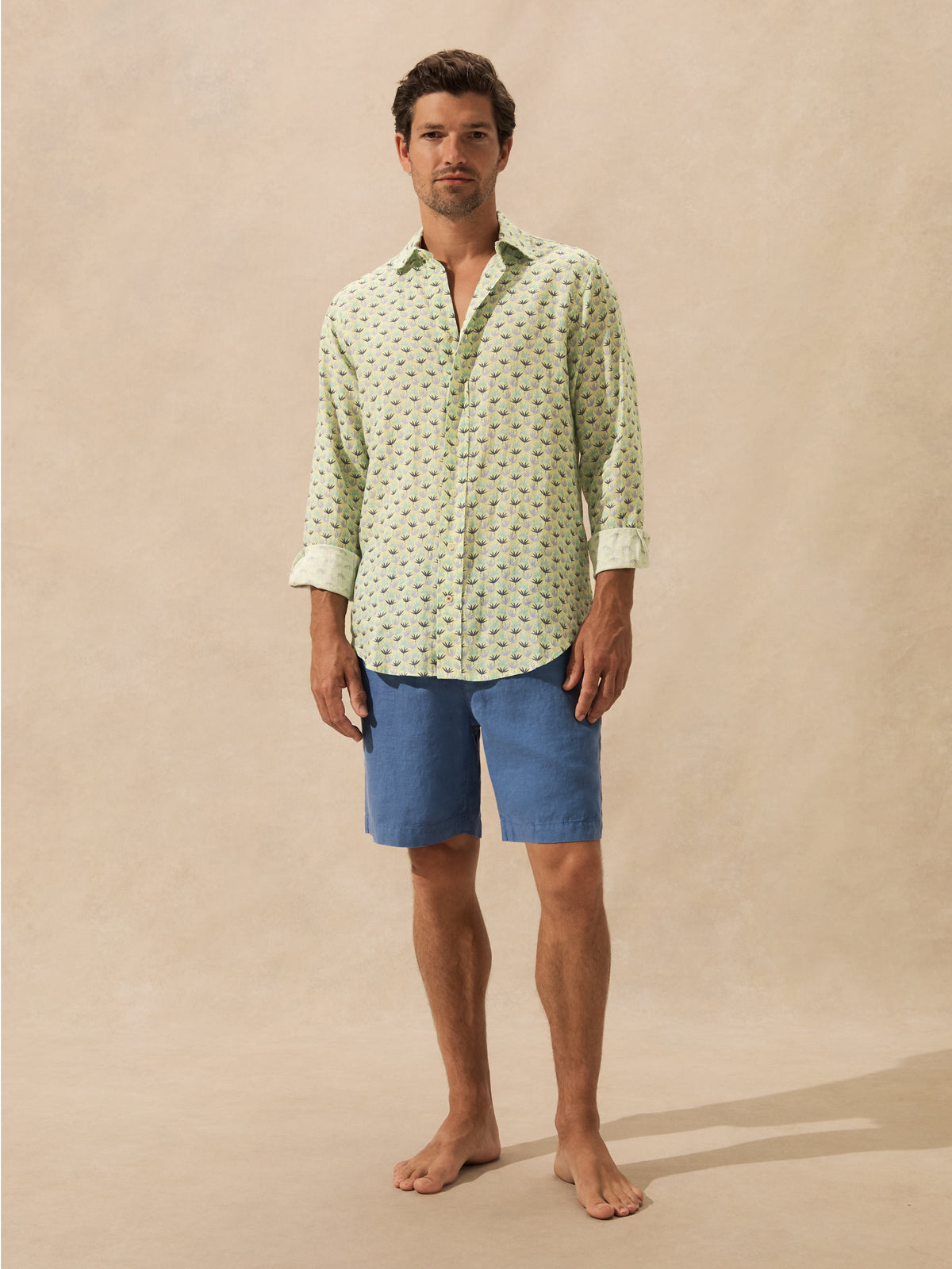 A man stands barefoot against a warm beige studio backdrop, wearing a light green long-sleeve button-up shirt with a small repeating dark print and blue shorts.