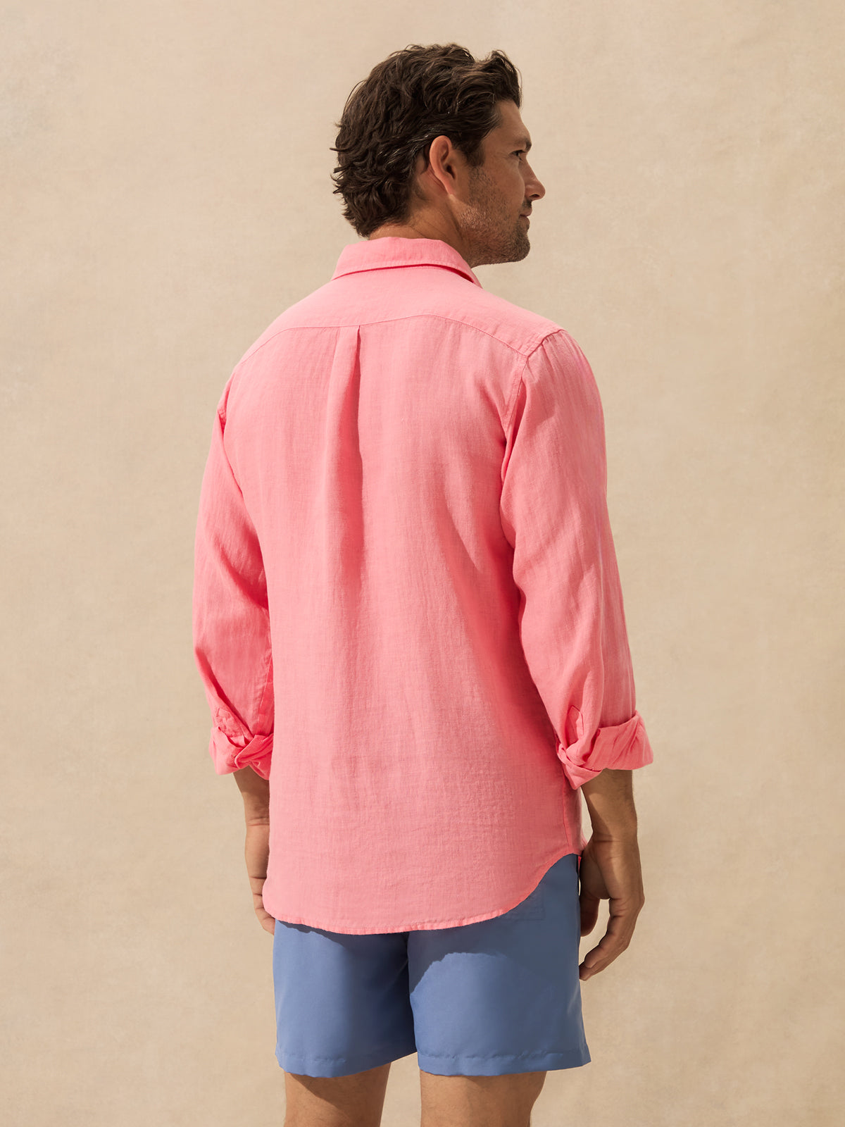 A man shown from the back wearing a pink long-sleeve linen shirt with rolled-up sleeves, standing against a textured wall.