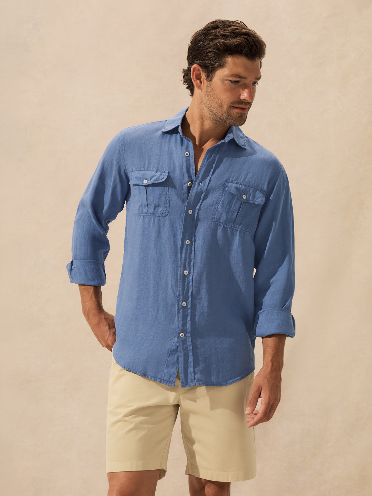 A man wearing a blue long-sleeve button-up shirt with two chest pockets and beige shorts, standing against a plain beige studio background.