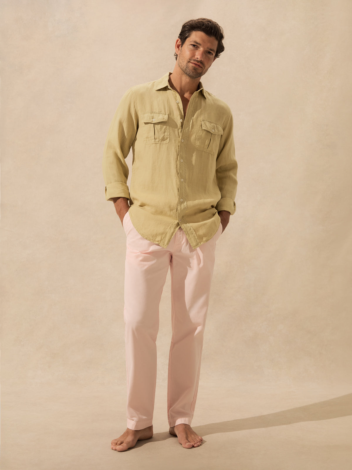 A man stands barefoot against a warm beige textured backdrop, wearing a khaki long-sleeve button-up shirt.