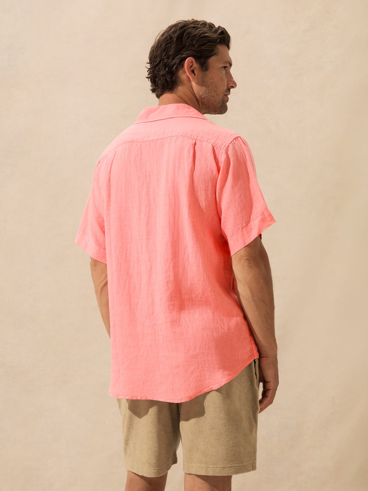 A man wearing a coral pink short-sleeve linen button-up shirt against a textured beige studio backdrop.