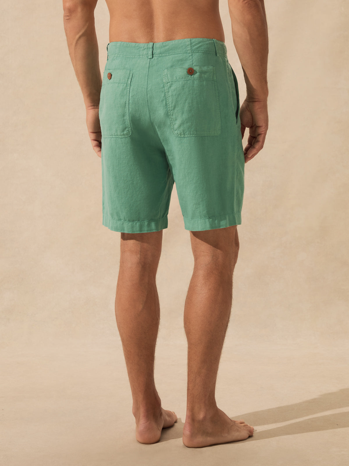 A man shown from the back wearing green linen shorts, standing barefoot against a warm beige textured background.