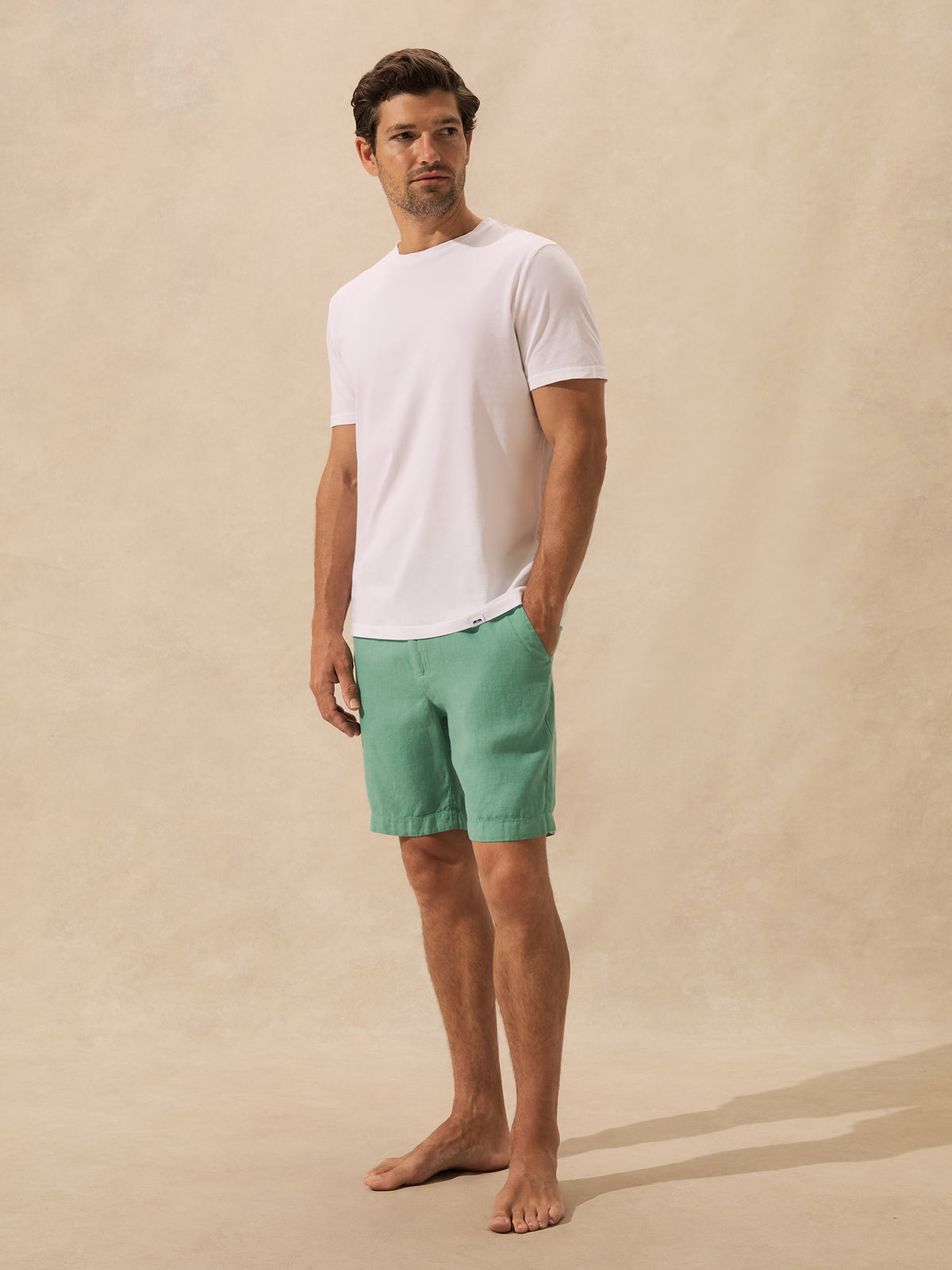 A man stands barefoot against a warm beige textured backdrop, wearing a plain white short-sleeve crew-neck t-shirt and green shorts.