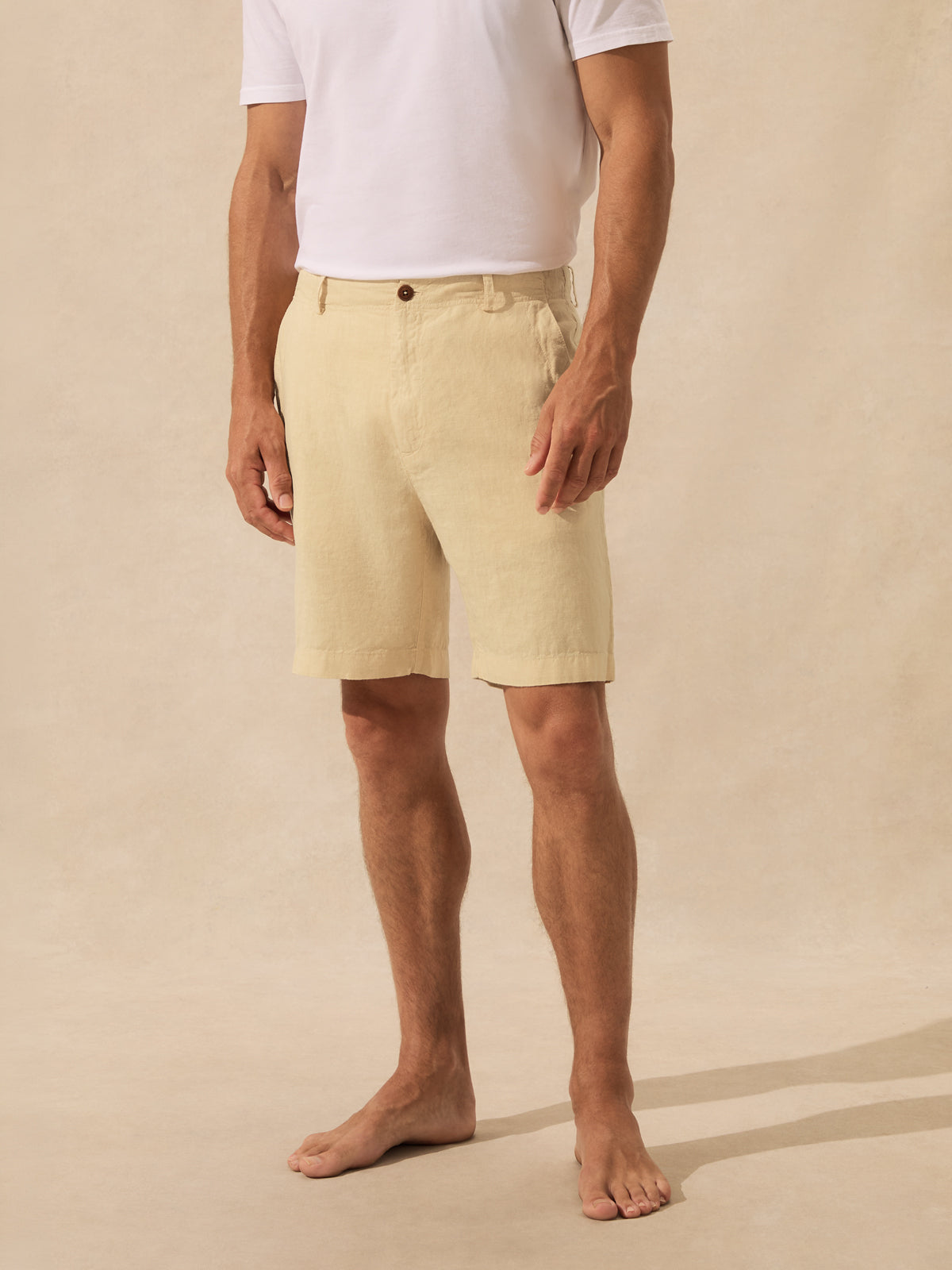 A man wearing light beige linen shorts and a white t-shirt, standing barefoot against a neutral beige studio background.