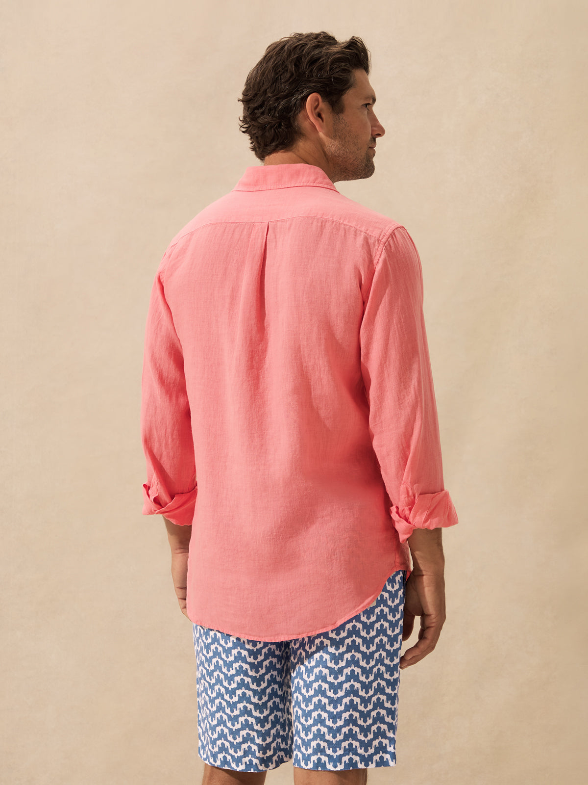 A man shown from the back wearing a coral pink long-sleeve linen shirt and blue patterned shorts against a warm beige studio backdrop.