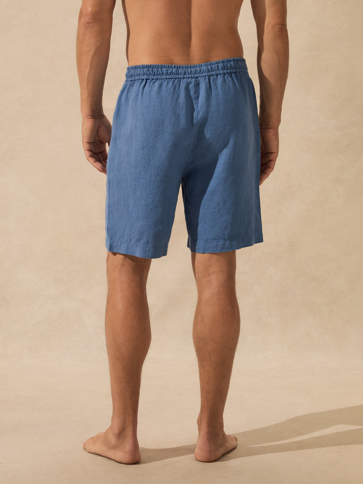 A man shown from the back wearing blue linen shorts, standing barefoot against a warm beige textured studio backdrop.