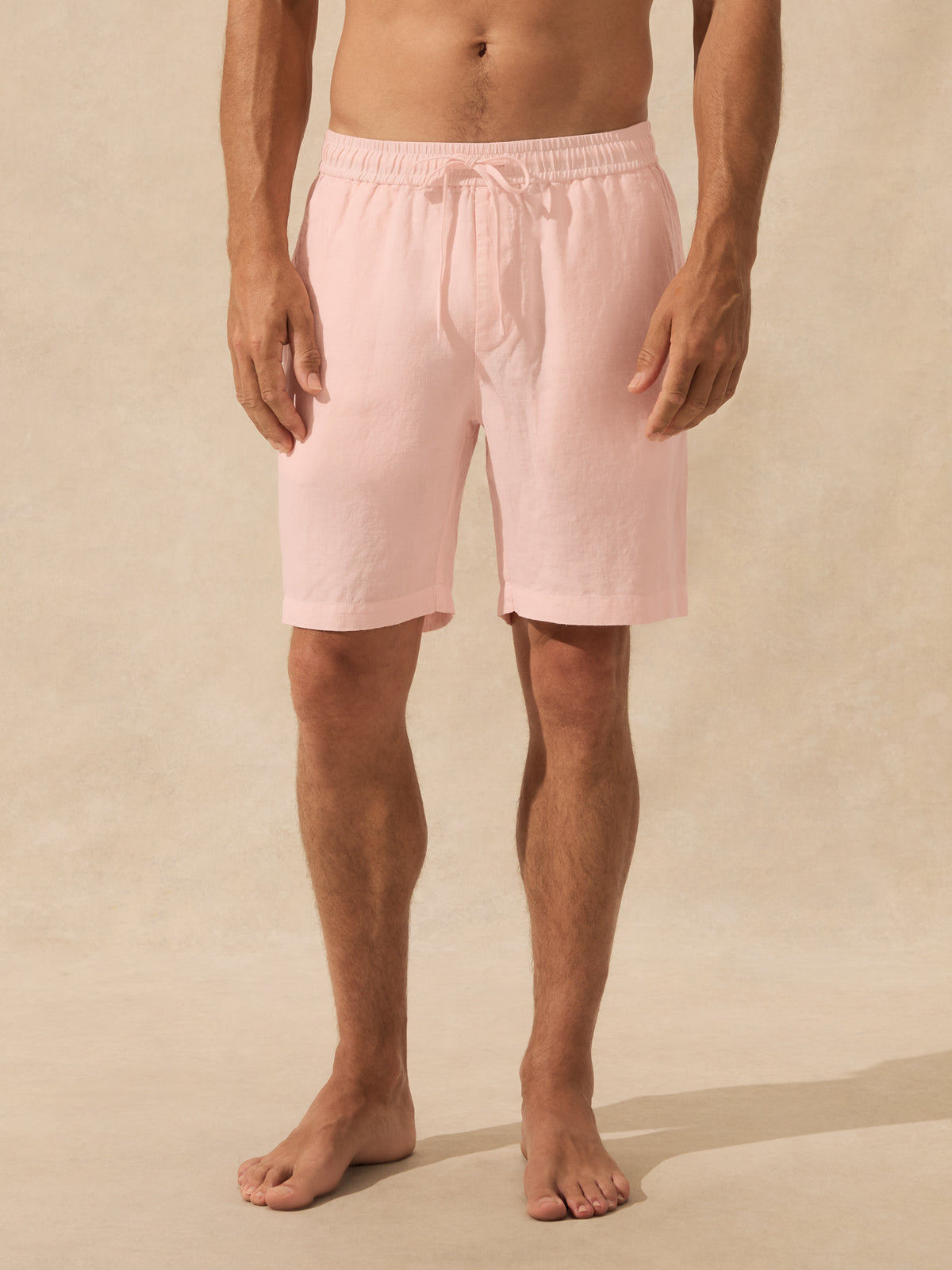 A man wearing light pastel pink drawstring shorts, shown from the waist down against a warm beige textured studio background.