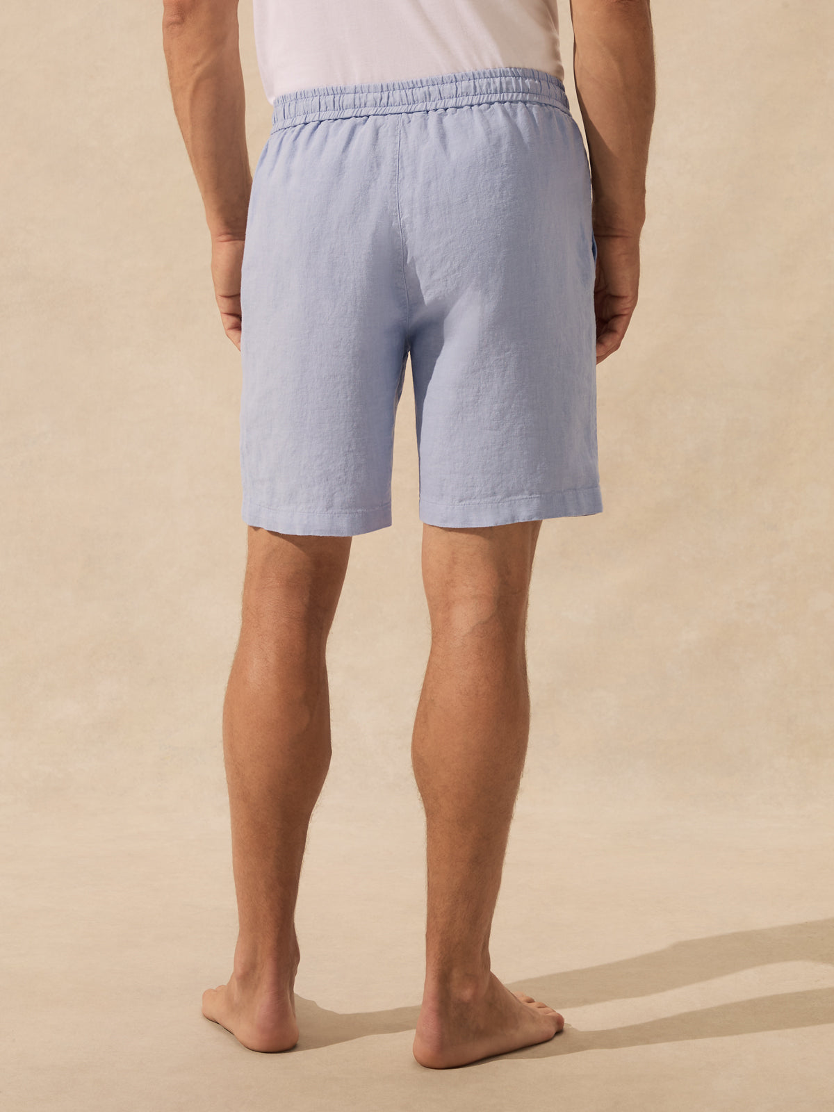 A man wearing light blue linen shorts, shown from the back against a warm beige textured background.