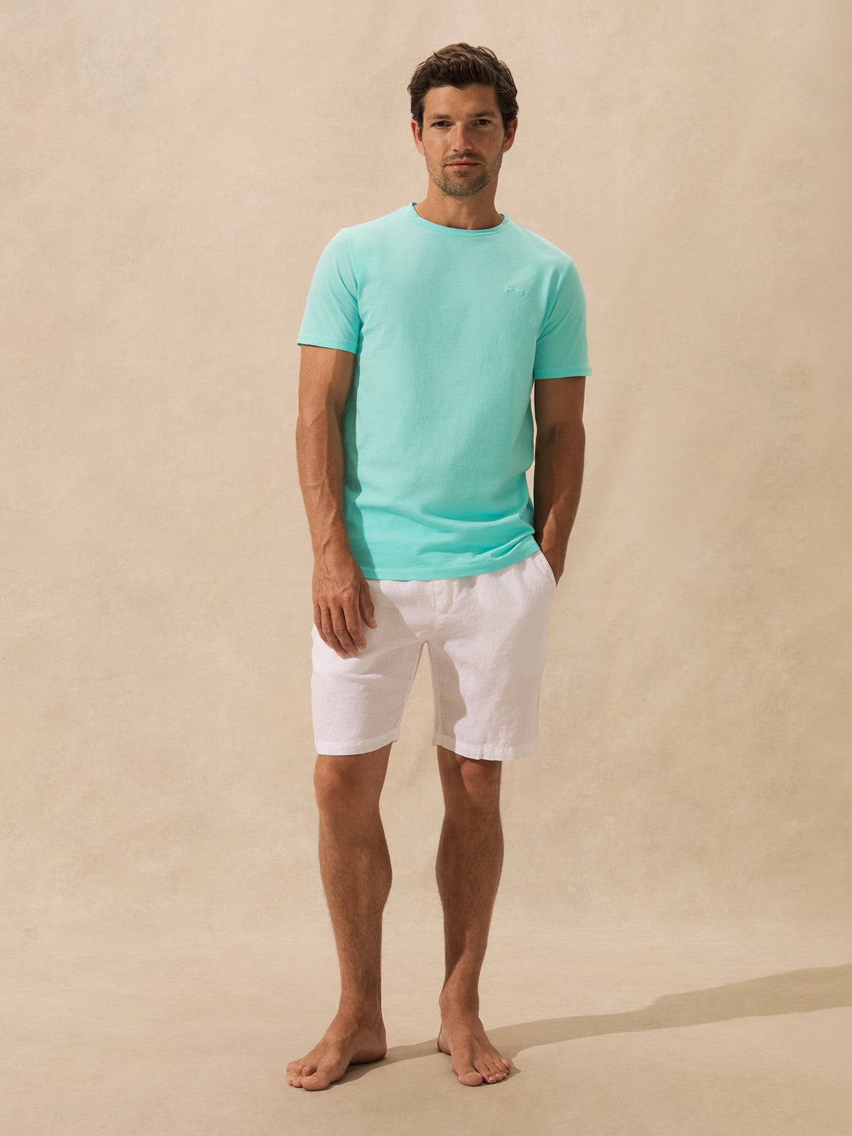 A man stands barefoot against a warm beige textured backdrop, wearing a light mint short-sleeve crewneck t-shirt and white shorts.