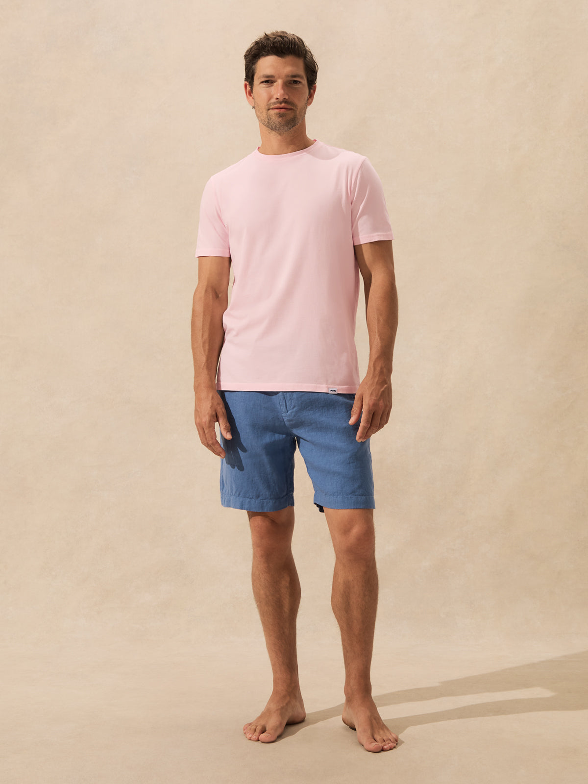 A man stands barefoot against a warm beige textured wall, wearing a light pink short-sleeve crewneck t-shirt and blue shorts.