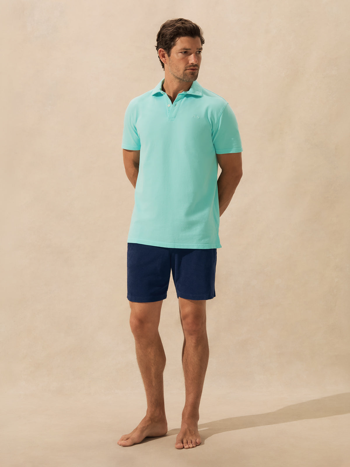 A man stands barefoot against a warm beige studio backdrop, wearing a light aqua green short-sleeve polo shirt and dark navy shorts.