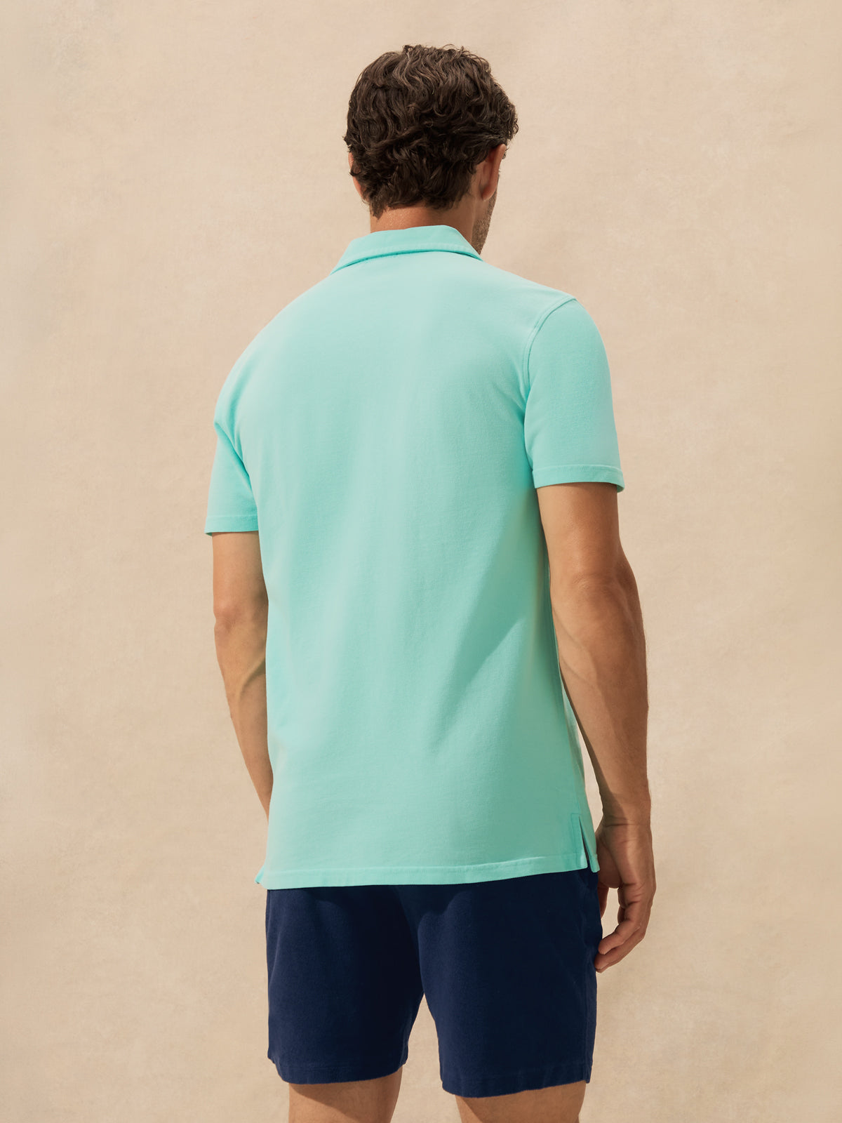 A man viewed from behind wearing a light mint green short-sleeve polo shirt and navy blue shorts, standing against a textured beige background.