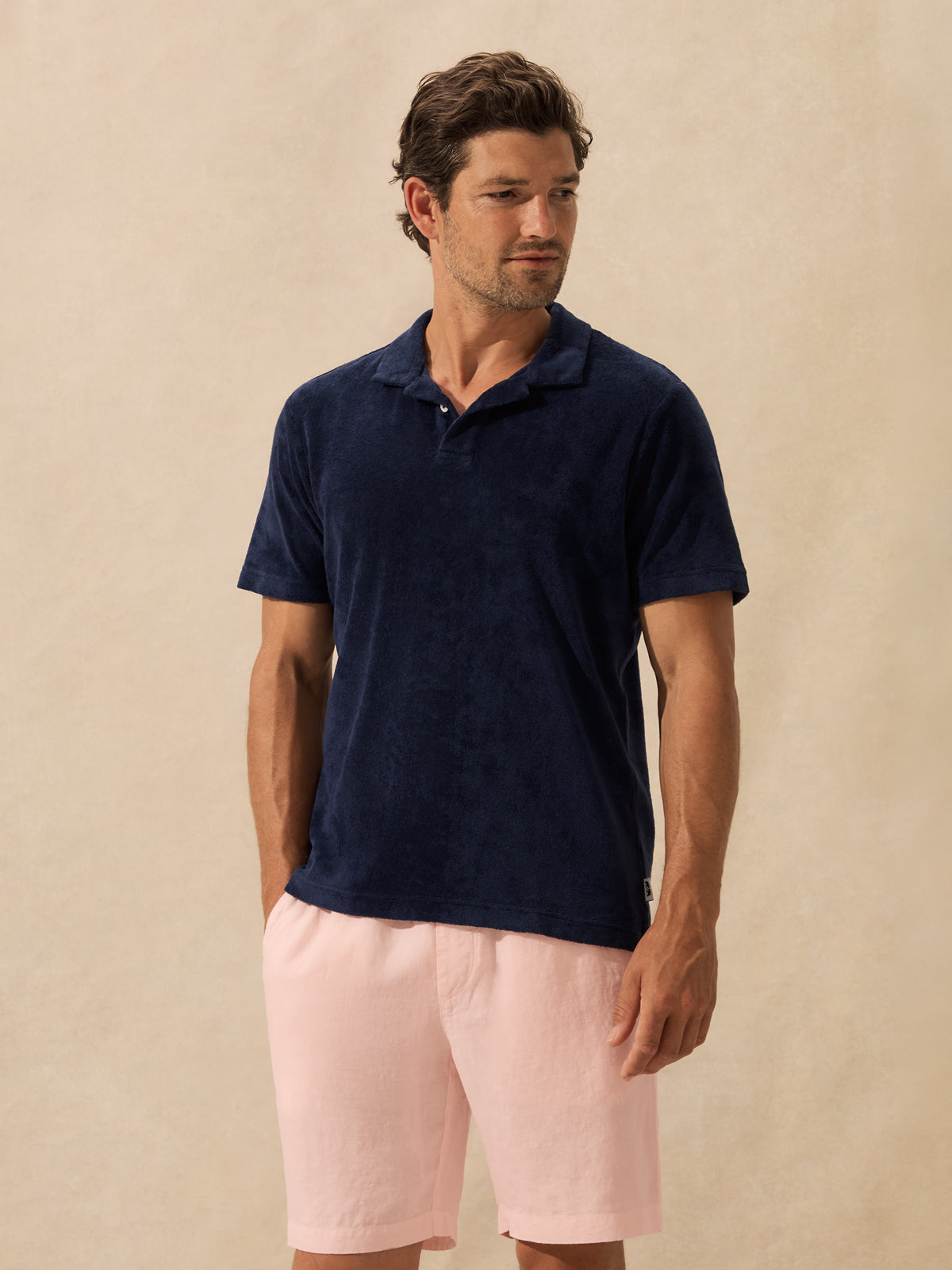 A man wearing a dark navy blue short-sleeve polo shirt and light pink shorts, standing against a textured beige background.