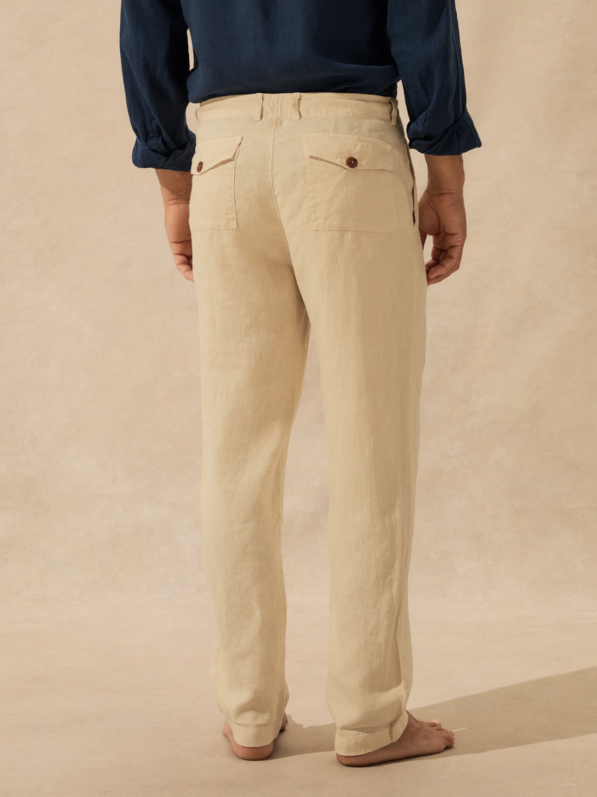 A person shown from the back wearing light beige linen trousers with two buttoned flap back pockets, paired with a navy blue shirt and bare feet, against a textured beige backdrop.