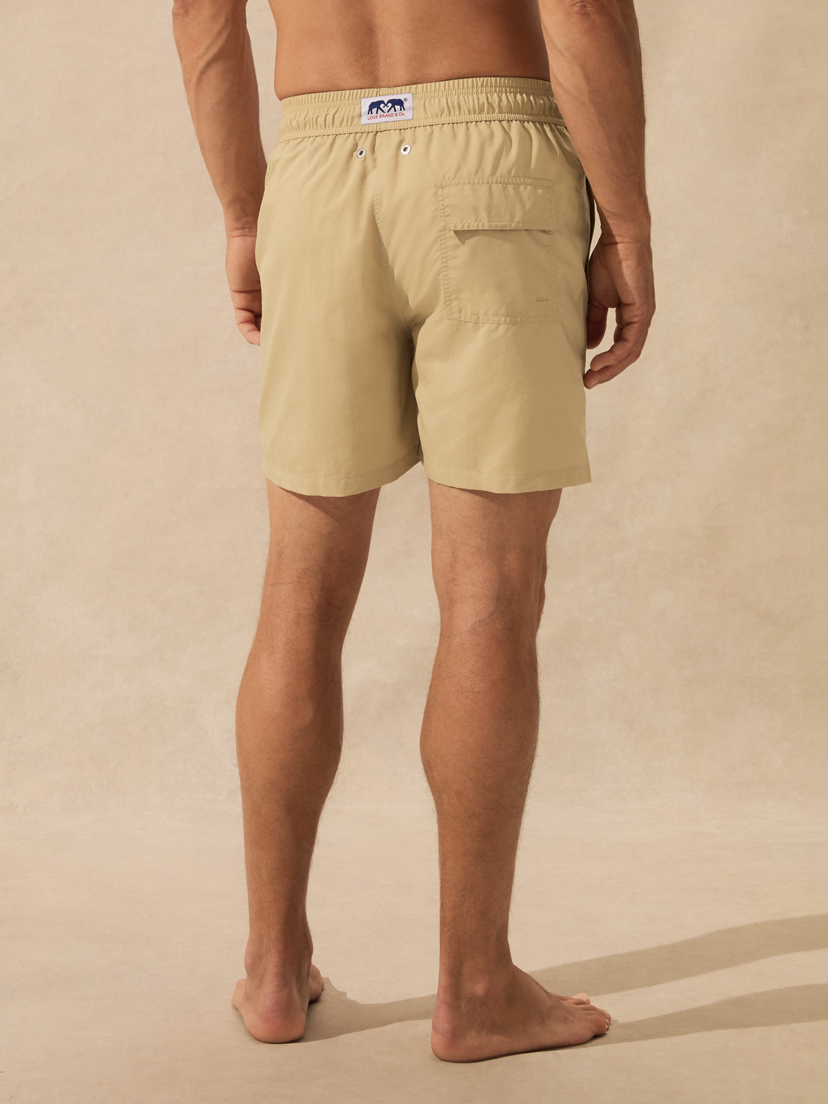 A man shown from the back wearing light beige swim shorts, standing barefoot against a textured beige studio backdrop.