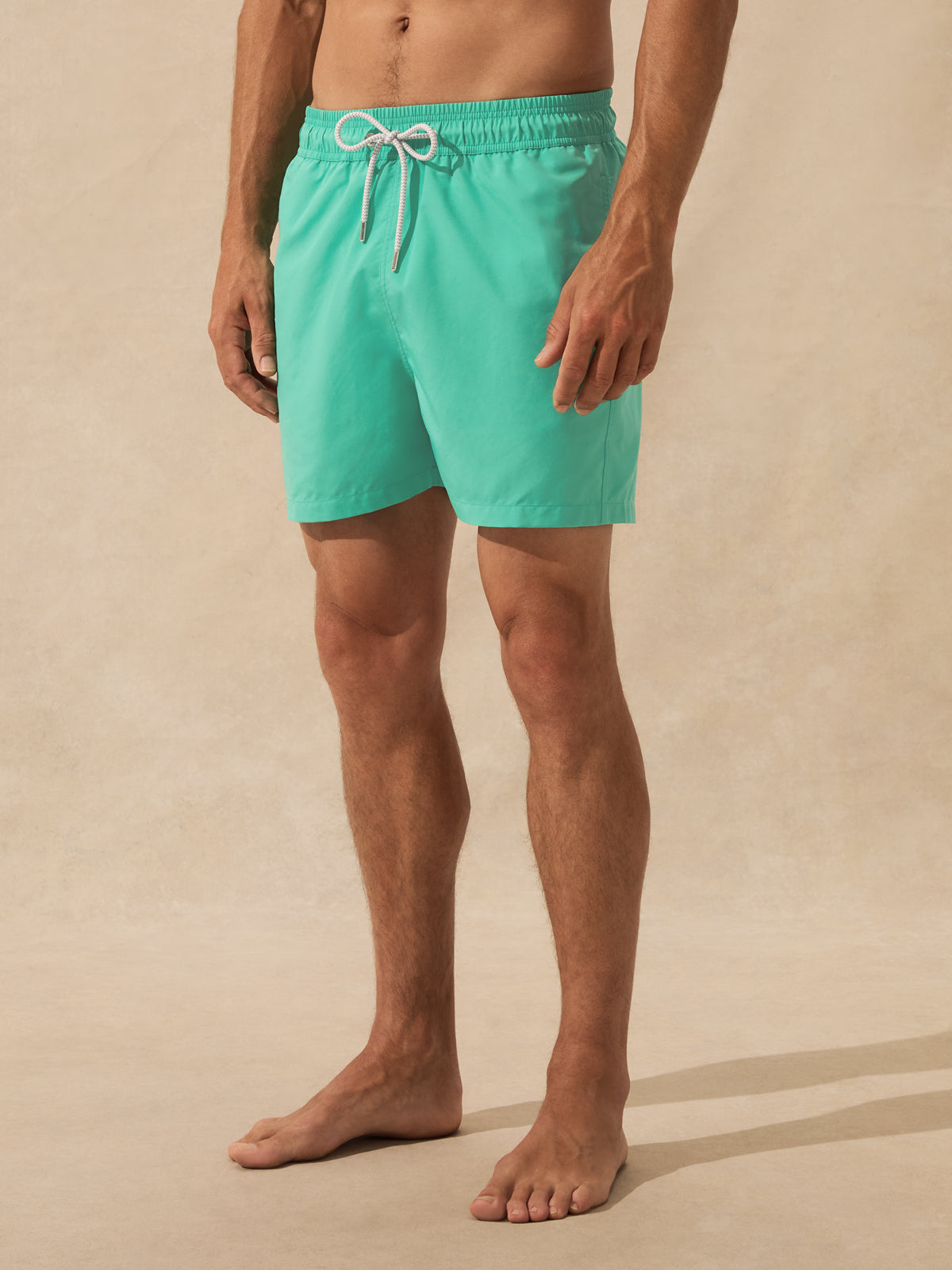 A man wearing bright turquoise swim trunks with a white drawstring, standing barefoot against a warm beige textured studio background.