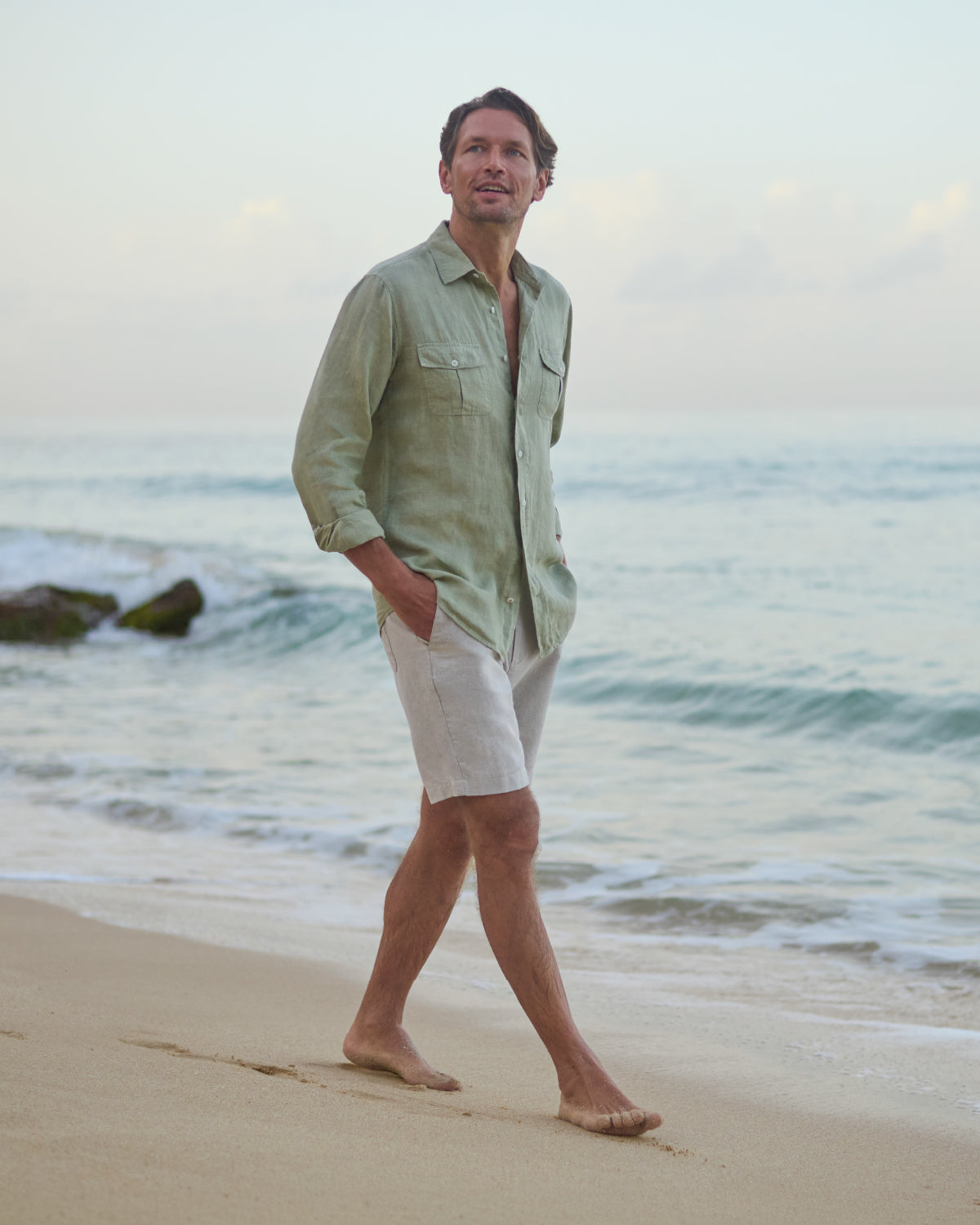 Man walking on a beach wearing a light green linen shirt and beige shorts.