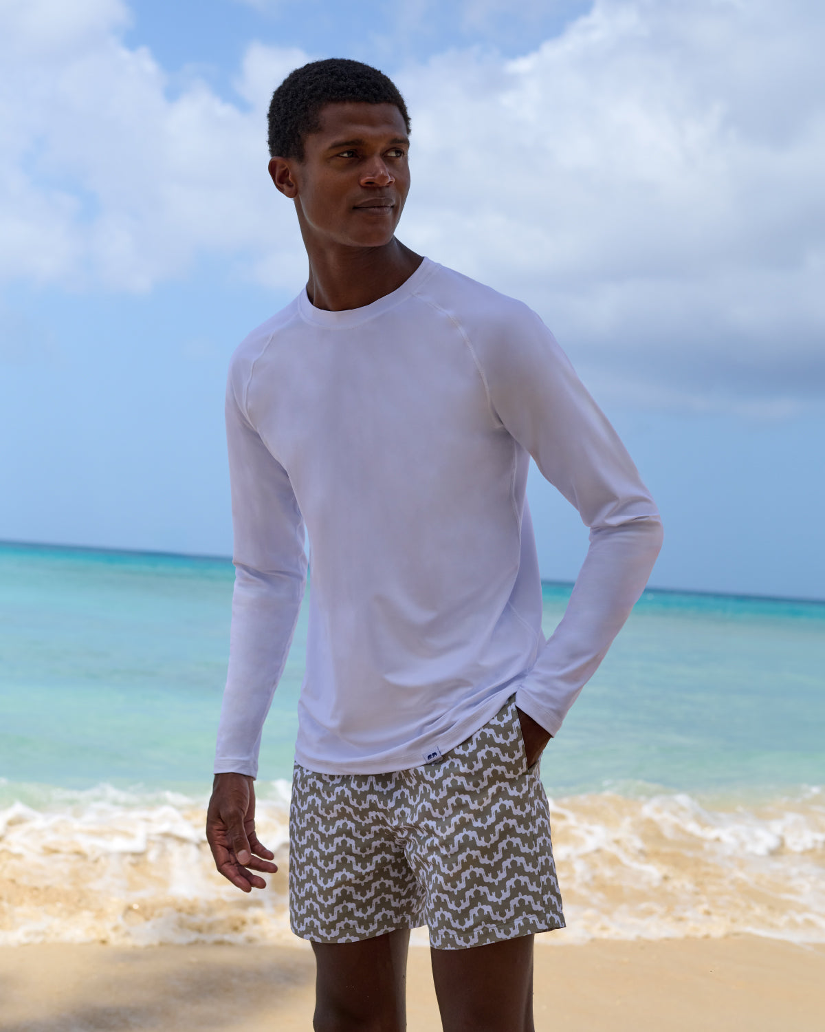 Man wearing a white long-sleeve rash vest and patterned swim trunks standing on a beach with ocean and sky in the background.