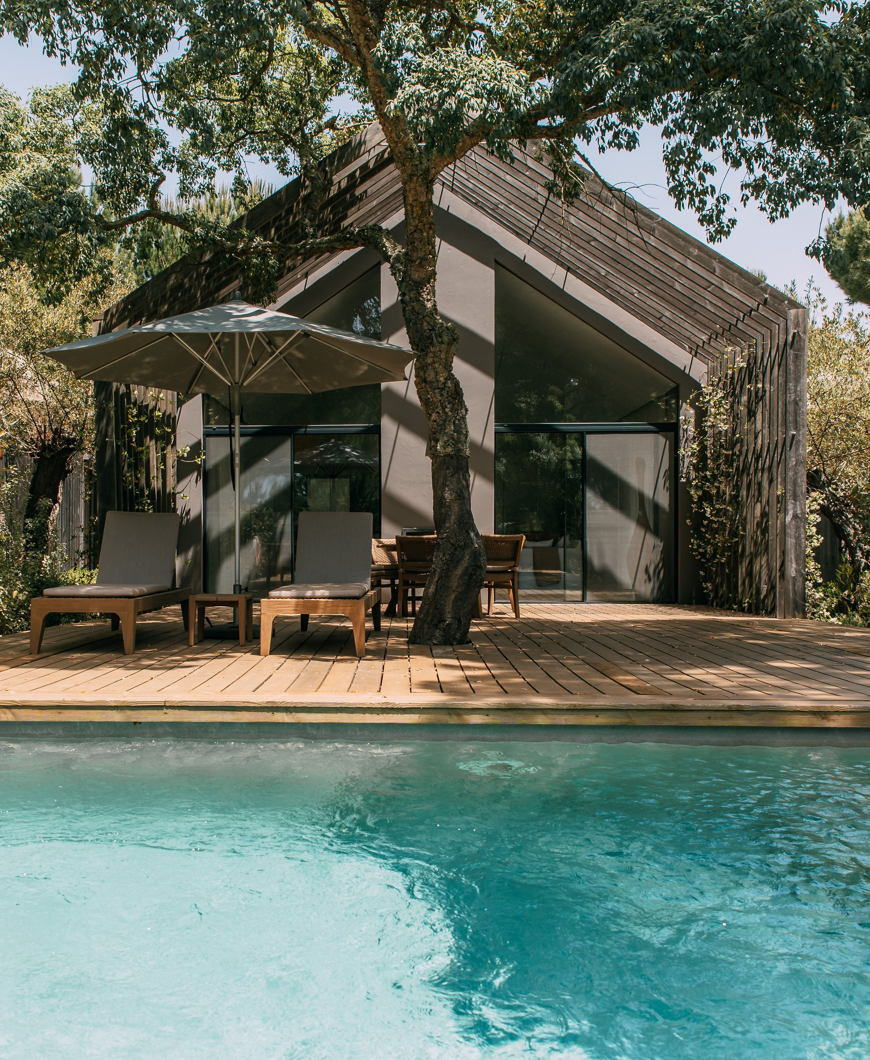A modern outdoor pavilion with a wooden deck, lounge chairs under a large umbrella, dining table, and a swimming pool in the foreground, surrounded by lush green trees.