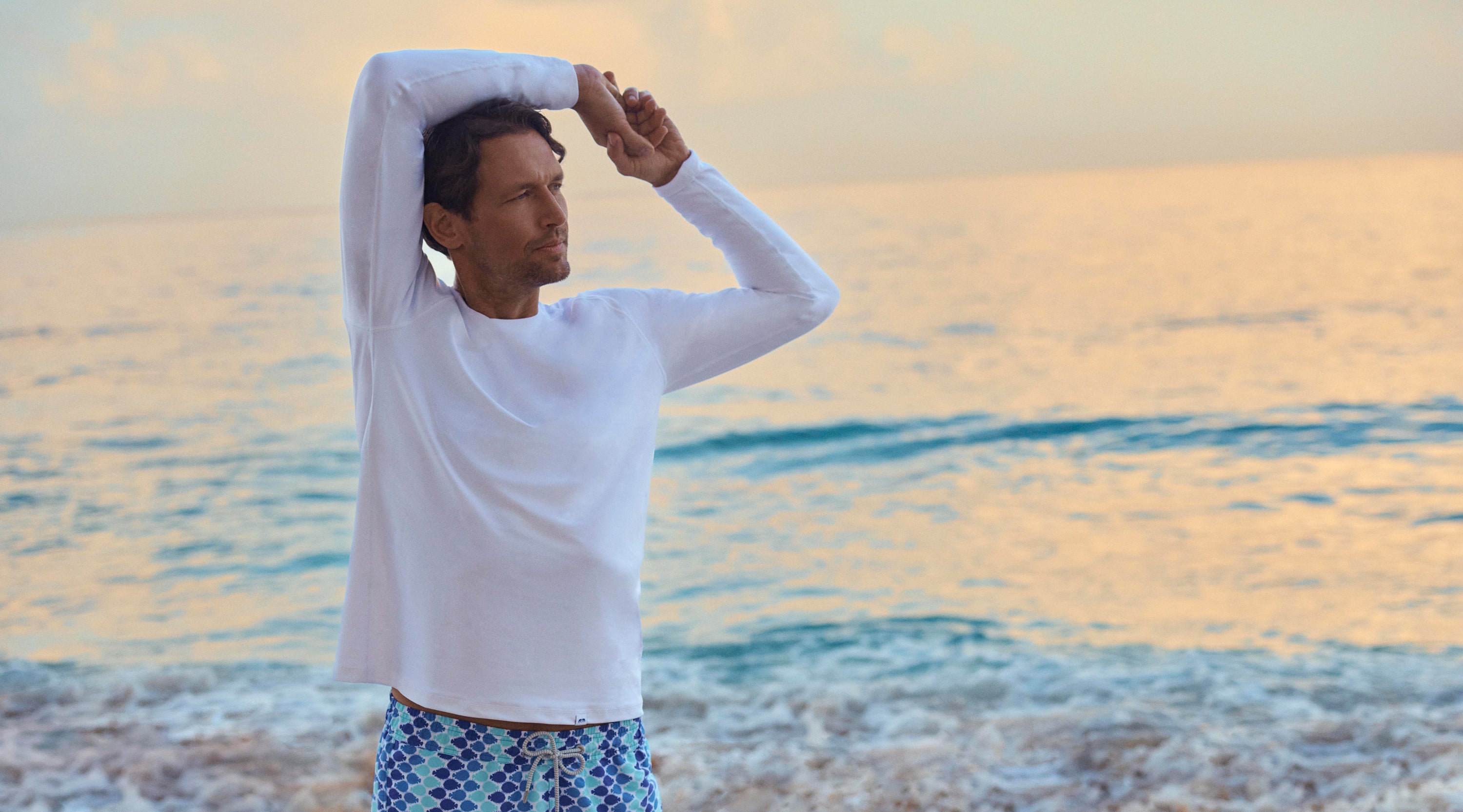 Man stretching on a beach with a sunset in the background wearing a white rash vest and swimming trunks