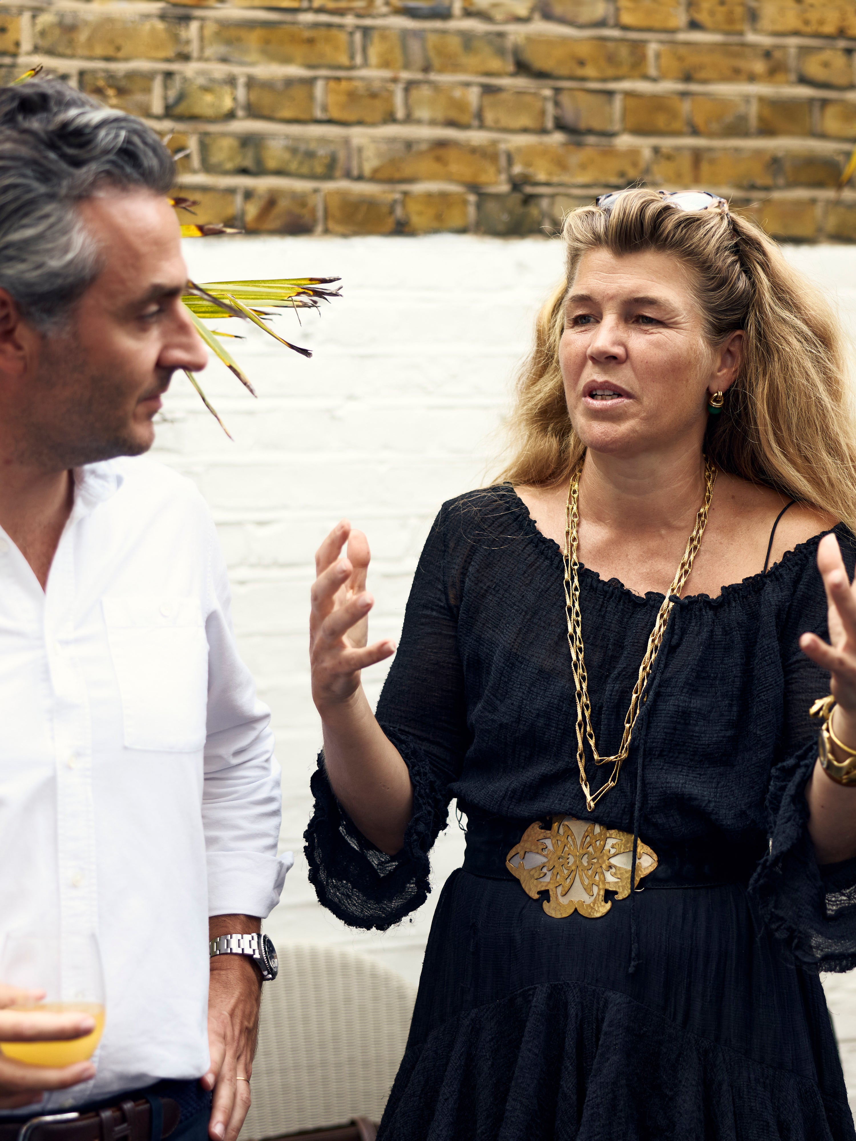 A woman in a black dress with a large gold belt buckle and layered gold necklaces is gesturing while speaking, standing next to a man in a white shirt in front of a brick and white wall background.