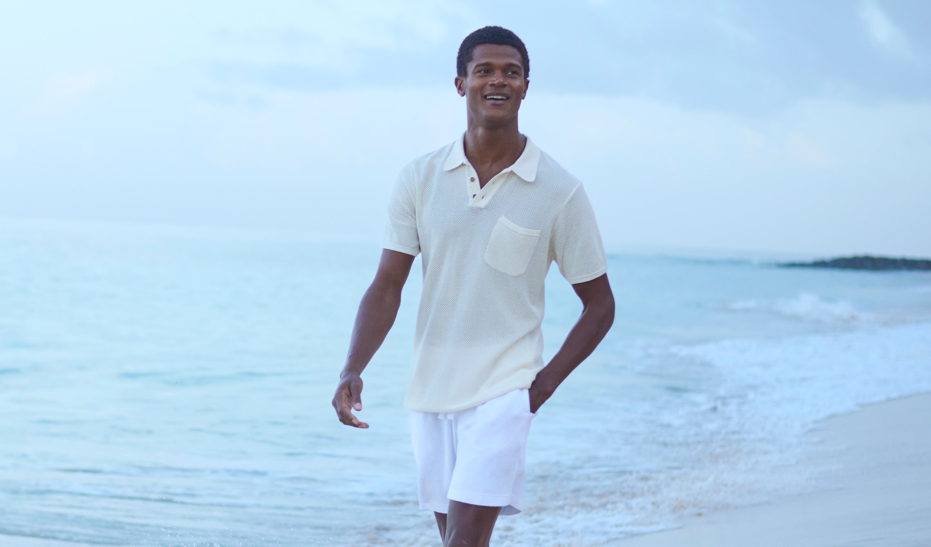 Man walking on a beach wearing a cream knitted polo shirt and white terry shorts.