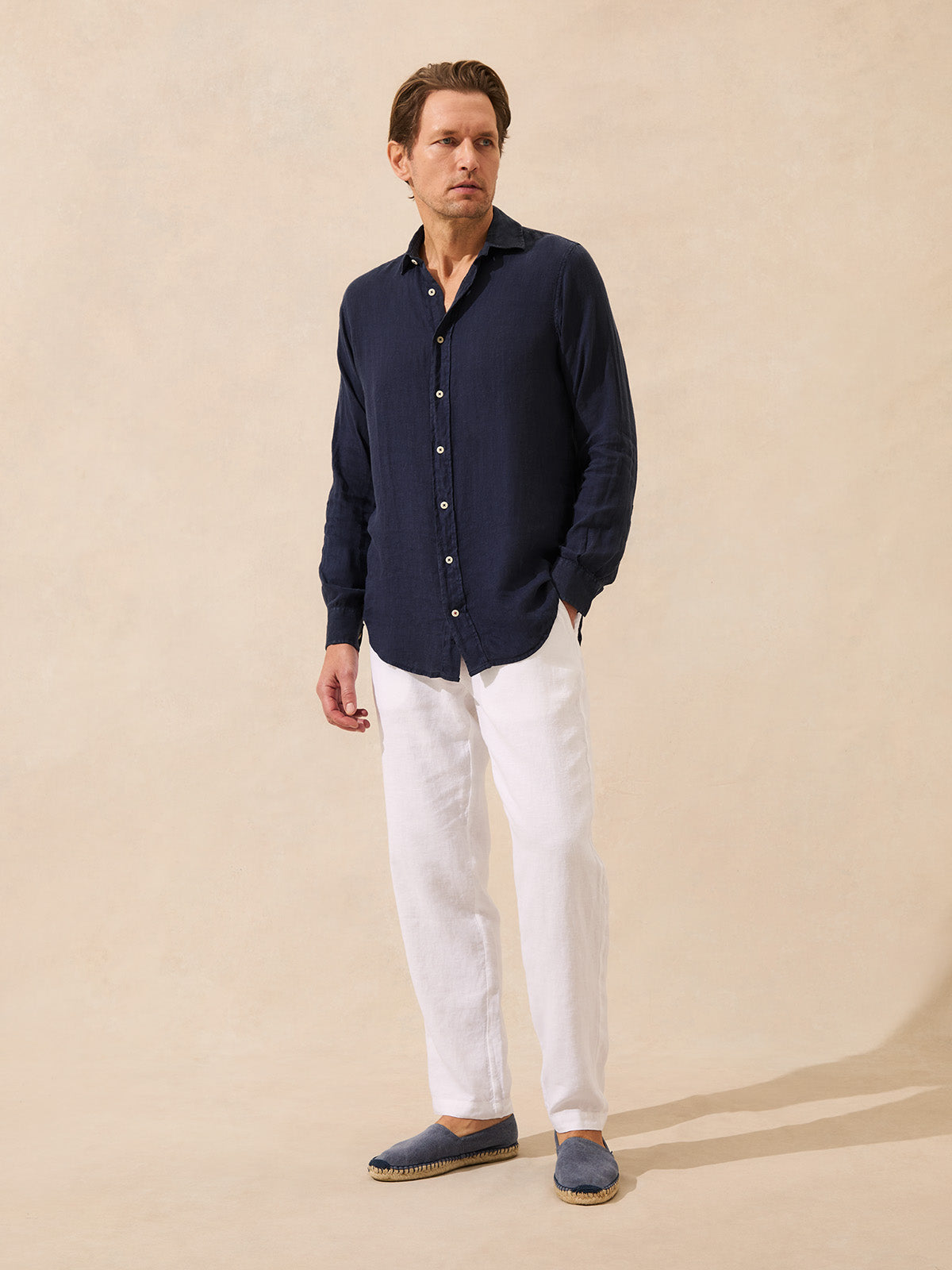 Man wearing a navy linen shirt and white linen trousers against a beige background