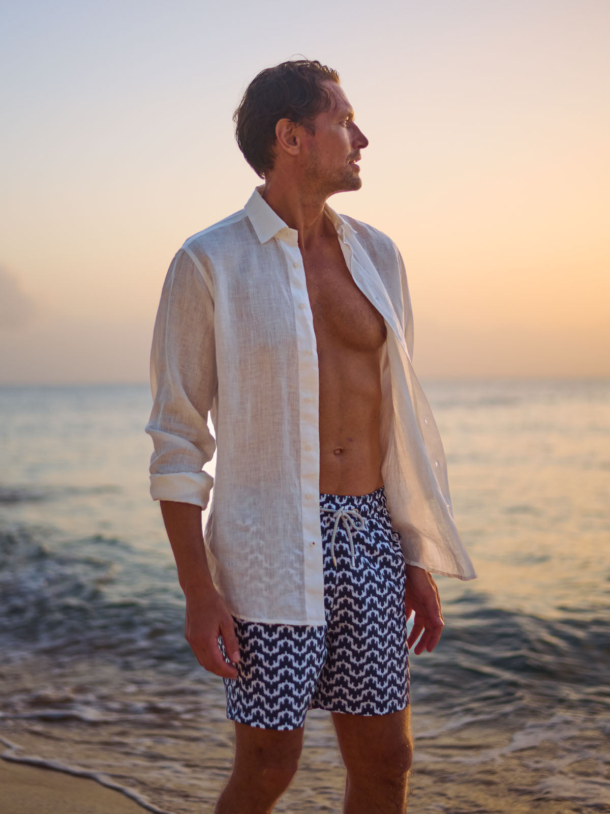 Man wearing a white linen shirt and patterned swim shorts standing on a beach at sunset.