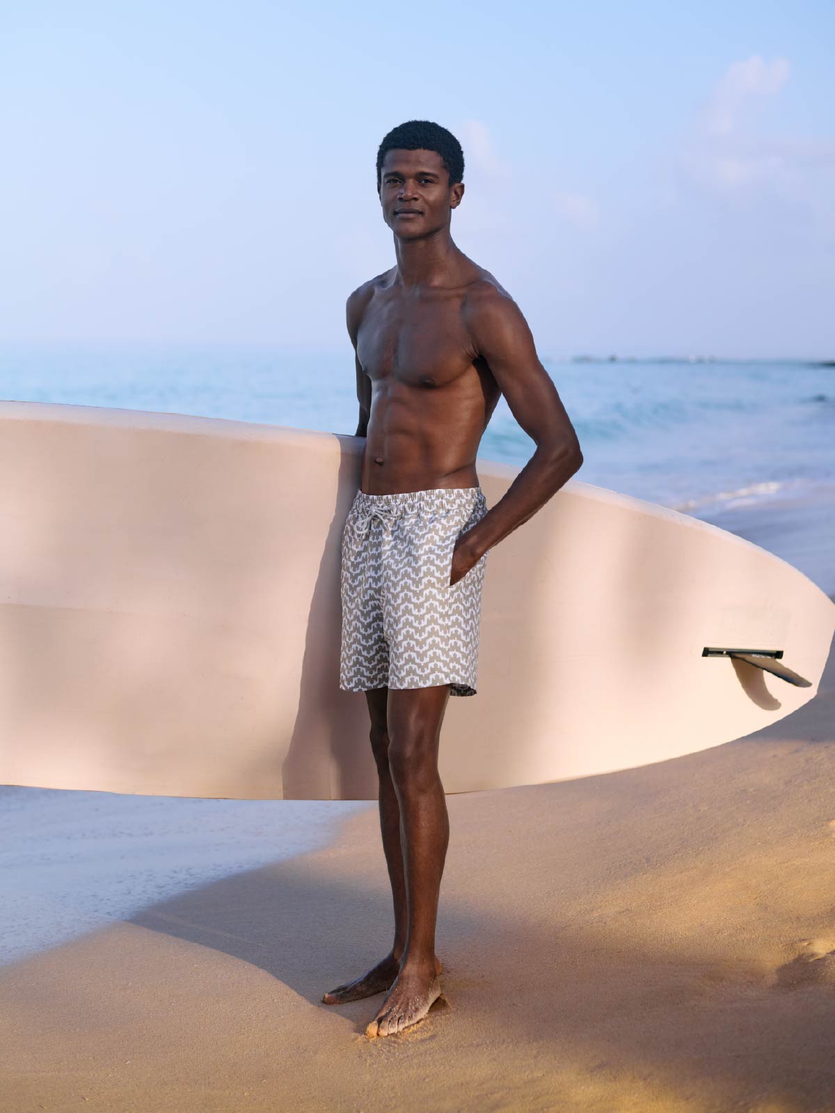 Man standing on a sandy beach with a surfboard, wearing patterned shorts.