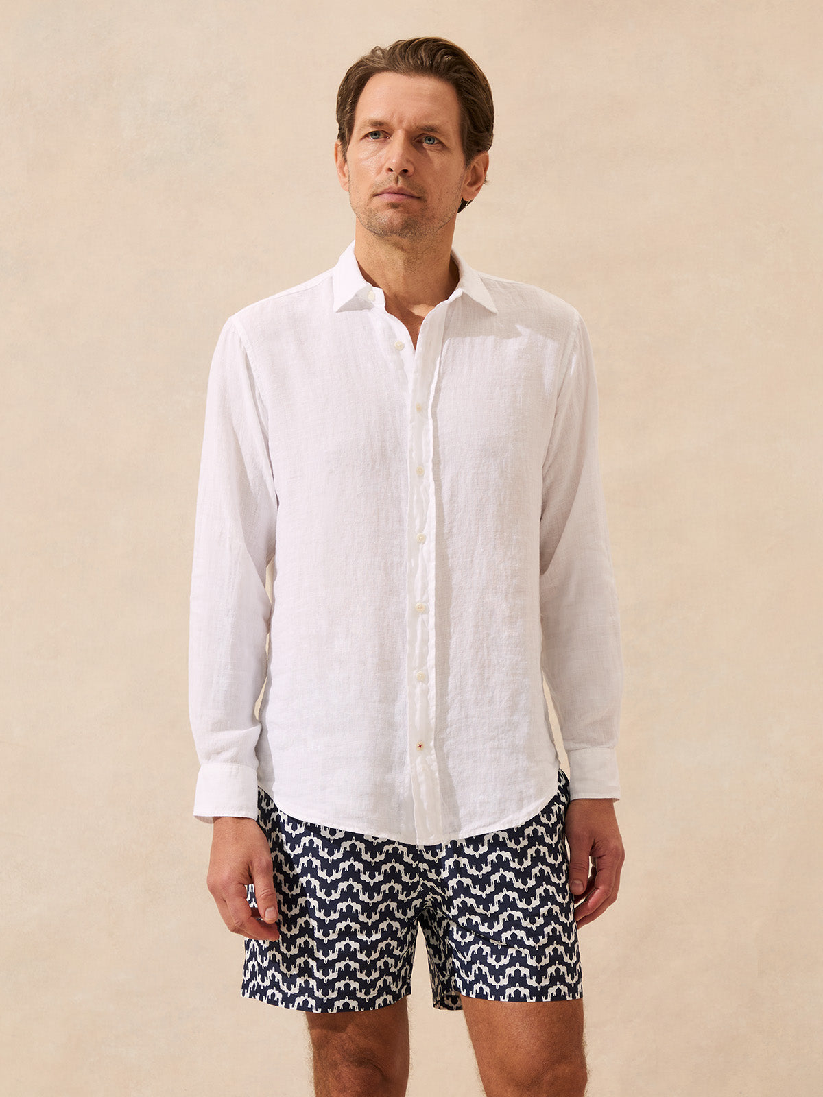 Man wearing a white linen shirt and patterned swim shorts on a beige background