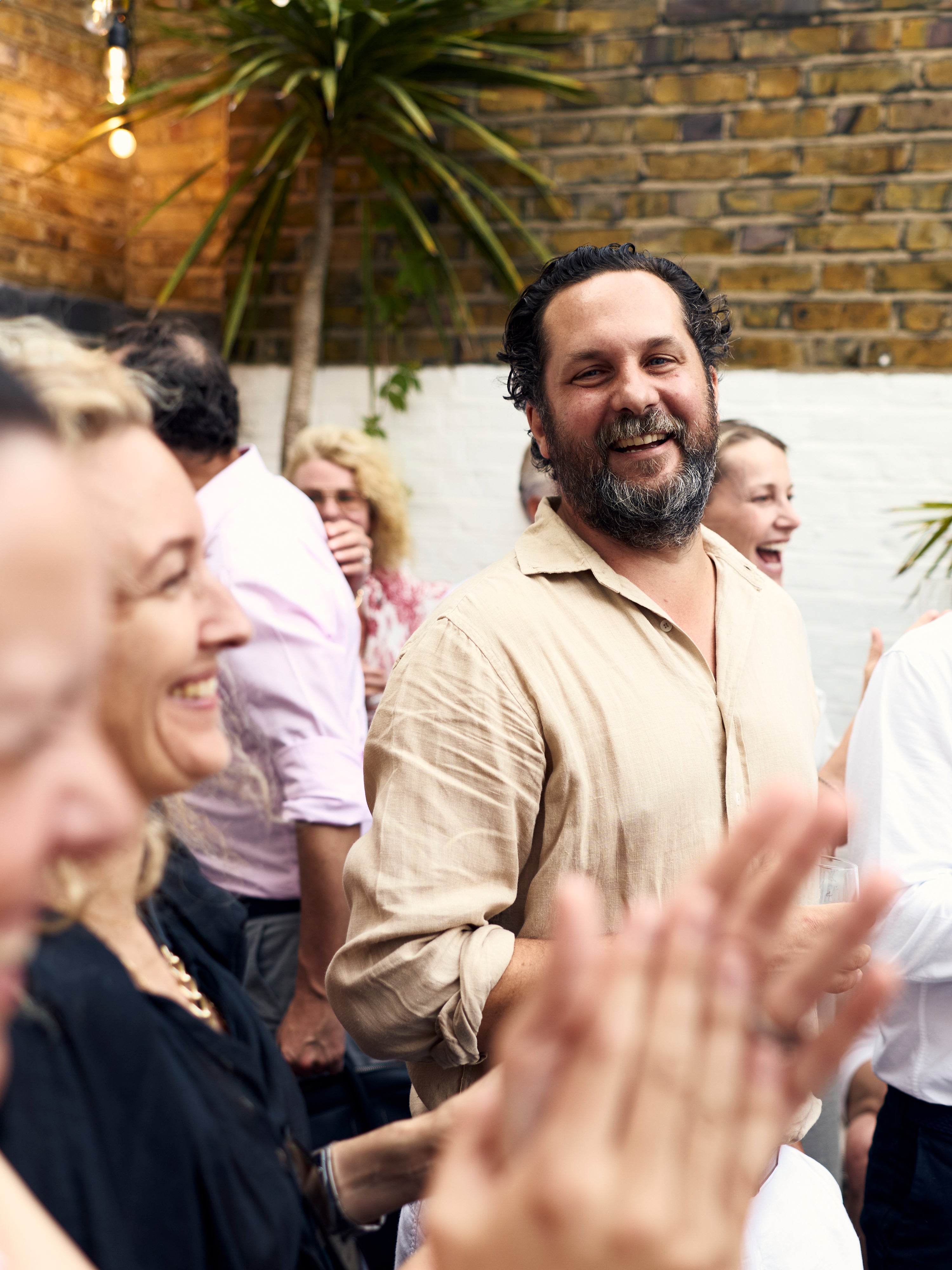 A smiling man in a light beige button-up shirt stands in a social setting with people clapping around him, against a brick wall background.