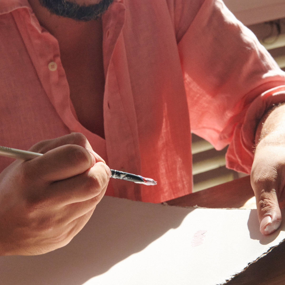 A close-up lifestyle shot of a person wearing a coral pink linen button-up shirt, holding a paintbrush and painting on a white canvas.