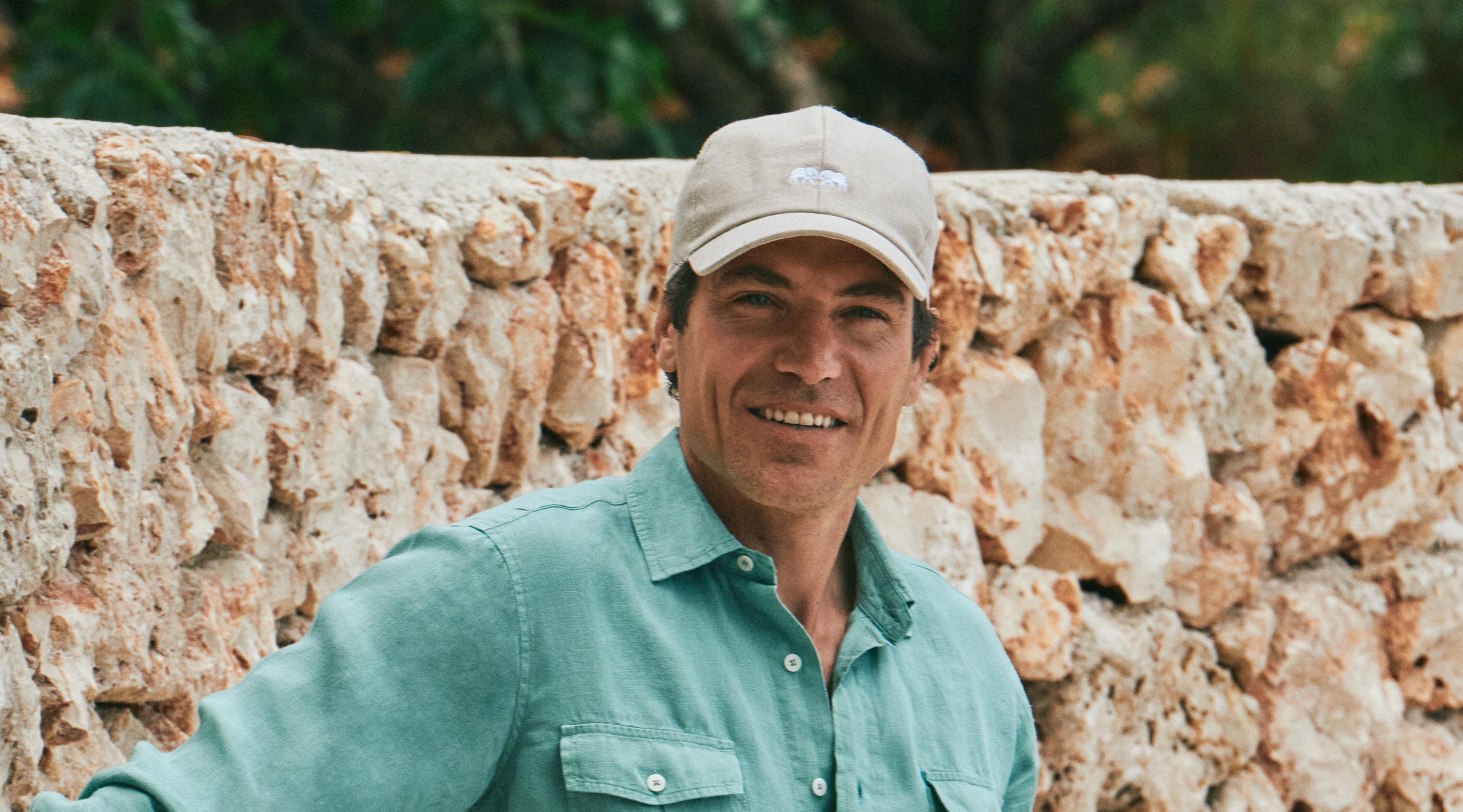 Man wearing a beige baseball cap and light green shirt standing in front of a stone wall with blurred greenery in the background.
