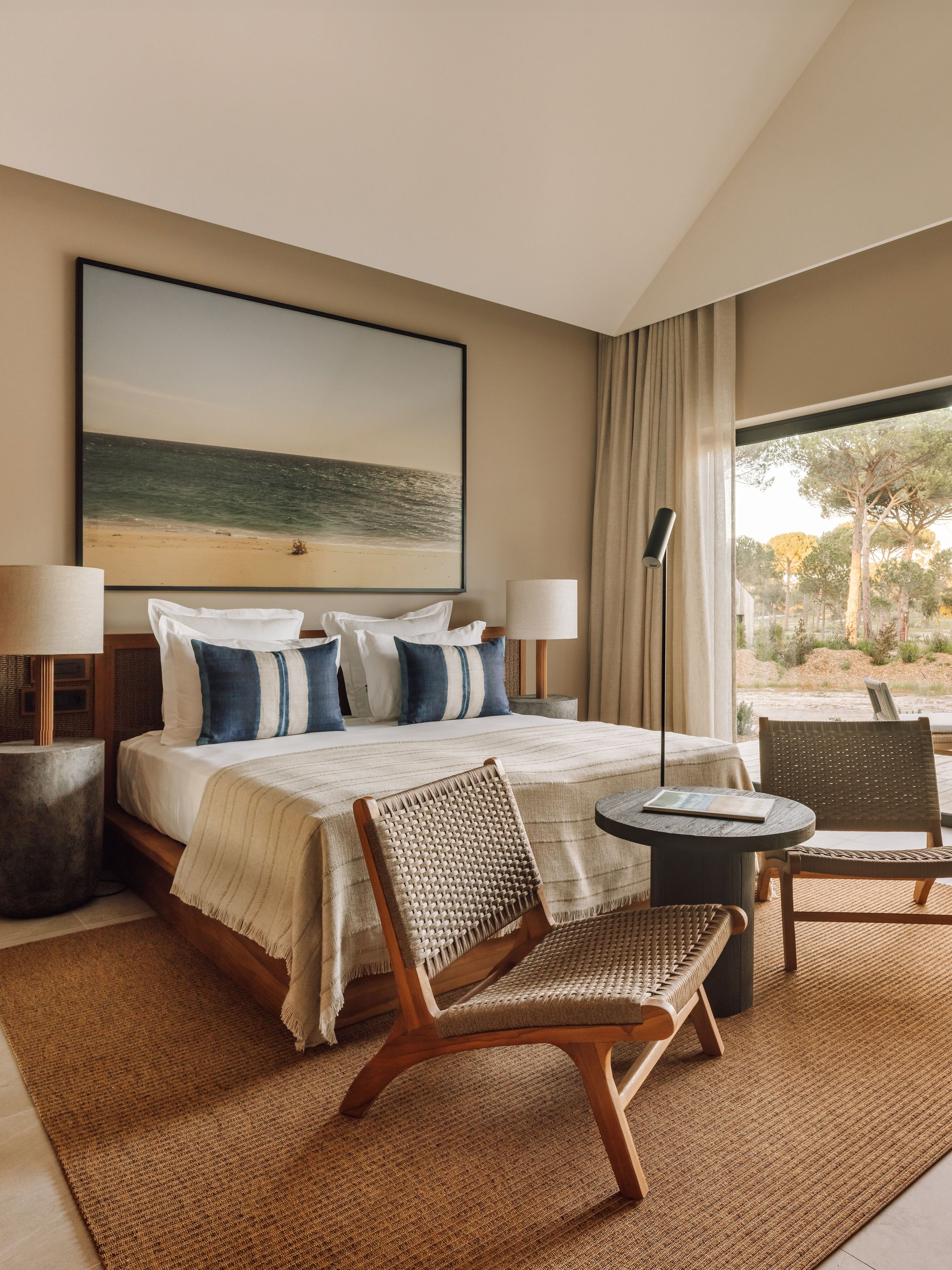 A styled bedroom interior featuring a bed with white bedding and blue striped pillows, a large framed beach landscape print on the wall, woven chairs, a round side table, and a window looking out onto a garden. The room is decorated in warm beige and brown tones.