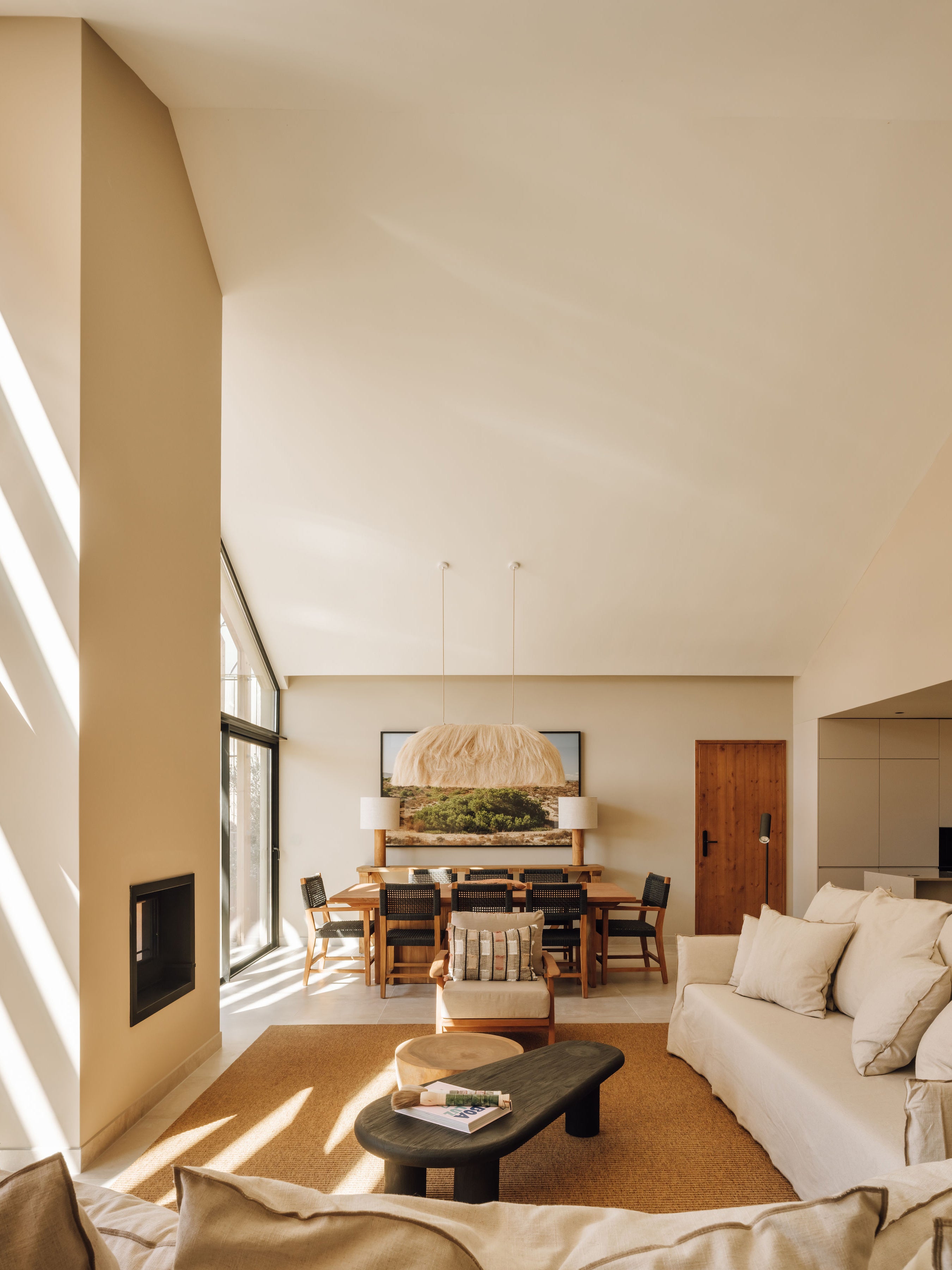 A modern, sunlit living room with a high vaulted ceiling, featuring a cream-colored sofa, a dark oval coffee table, a woven rug, and a dining area in the background.