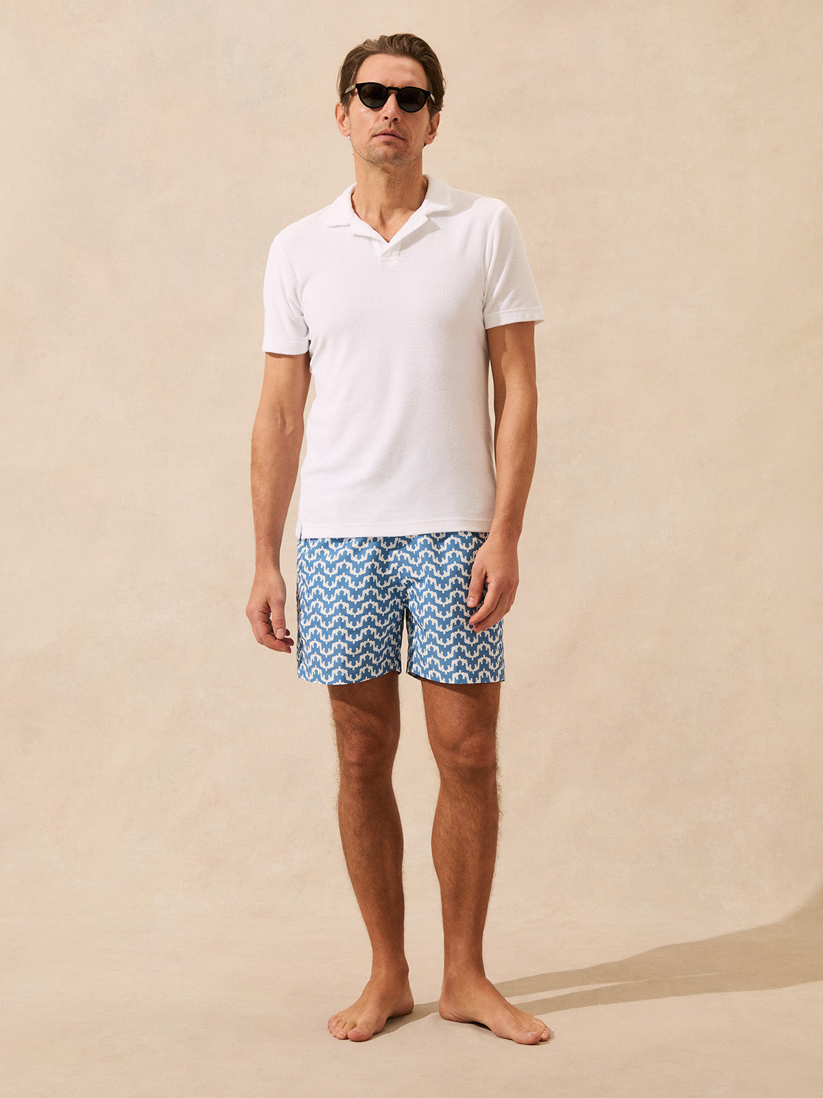 Man wearing a white terry polo shirt and blue patterned swim shorts on a beige background