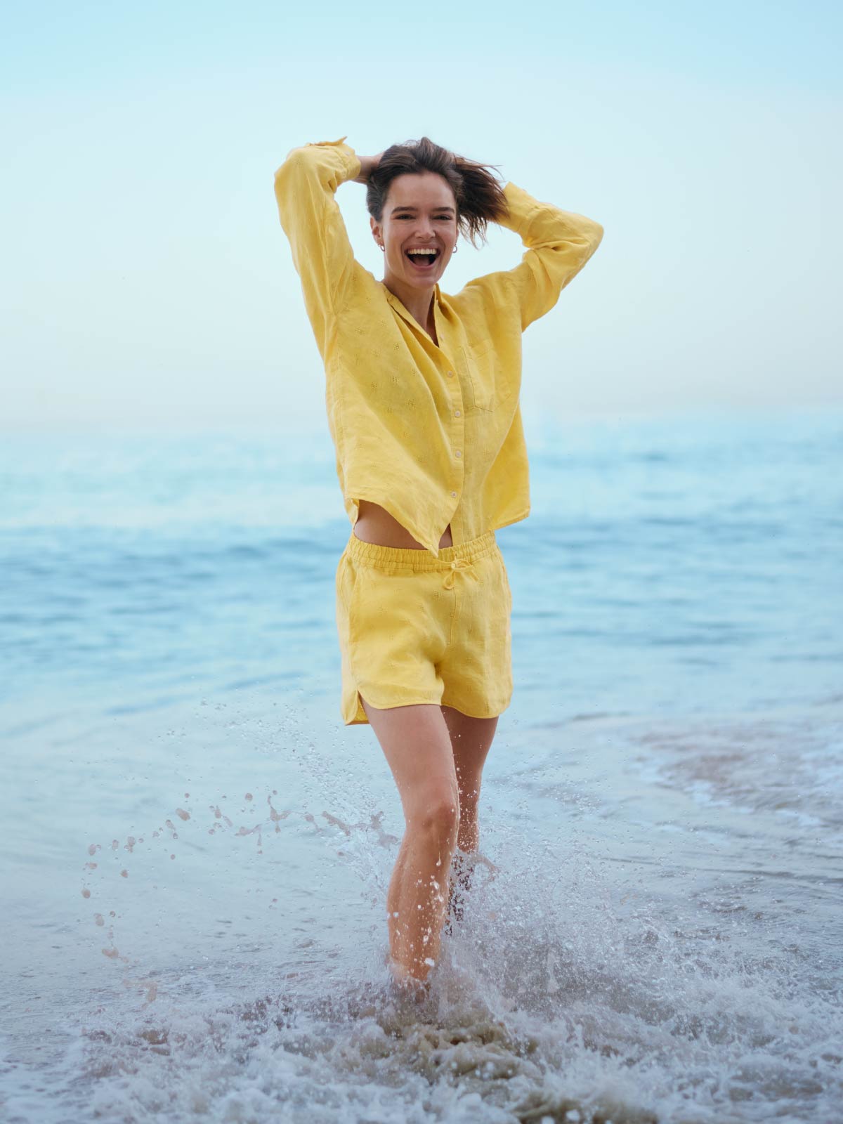 Woman wearing a yellow linen outfit standing in shallow water at the beach.