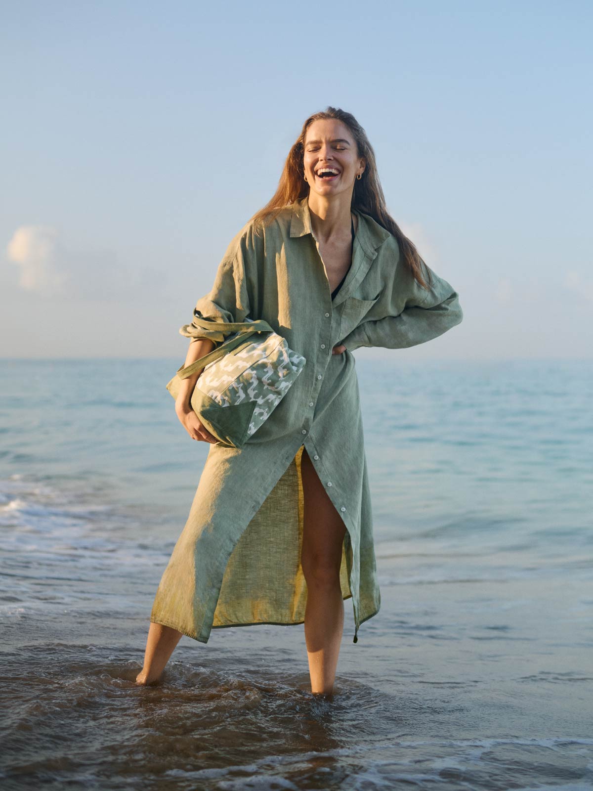 Woman in a long green shirt dress standing in shallow water at the beach.