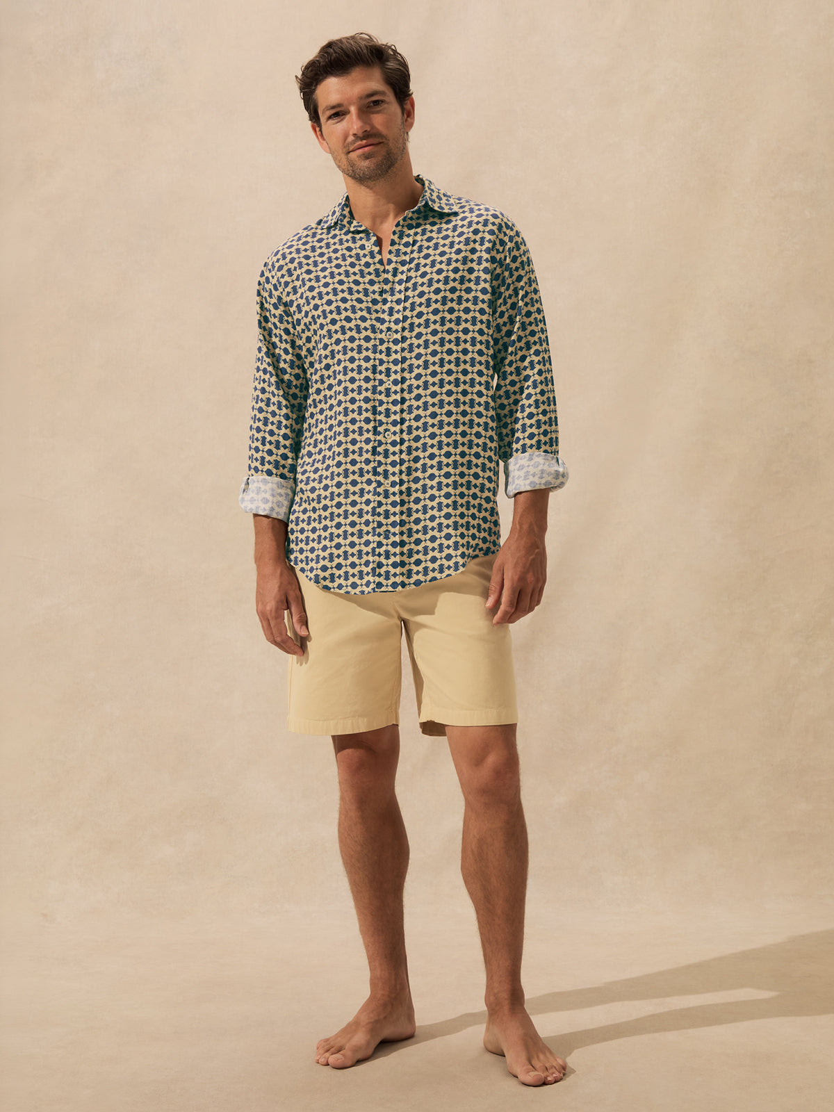 A man stands barefoot against a warm beige textured backdrop, wearing a long-sleeve button-up shirt with a brown and blue geometric pattern and light beige shorts.