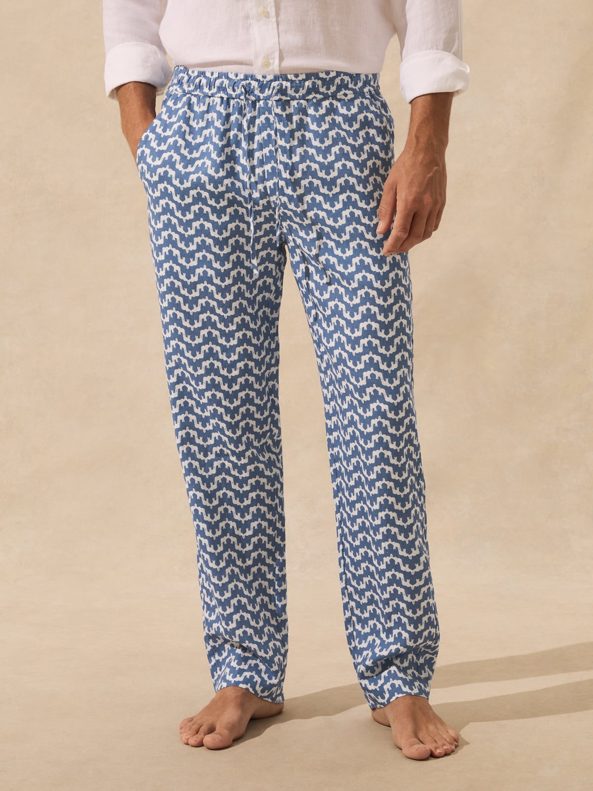 A person wearing blue and white patterned lounge pants with a white shirt, standing barefoot against a beige studio background.