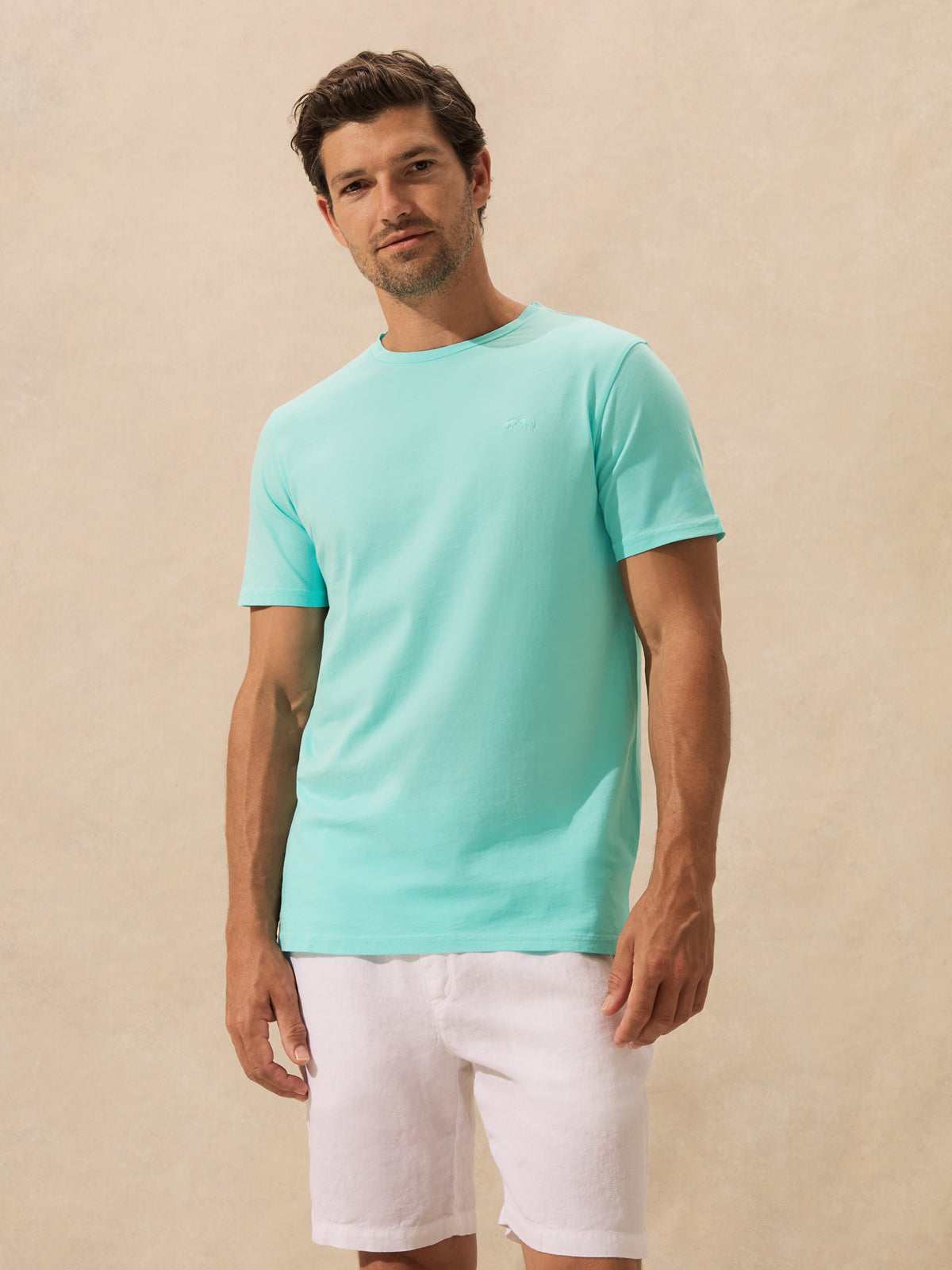 A man wearing a light mint green crew-neck t-shirt and white shorts, standing against a textured beige wall.