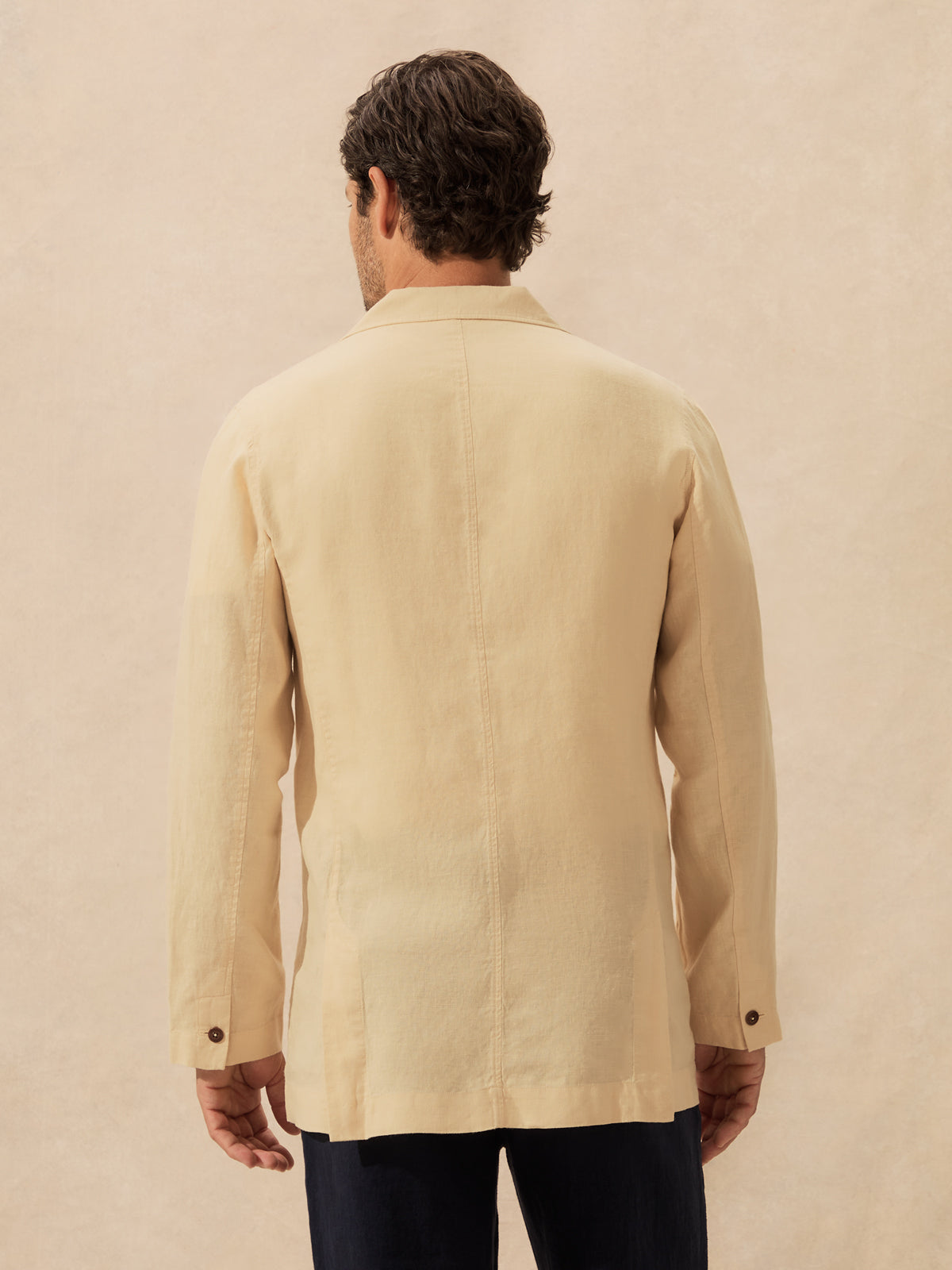 A man viewed from behind wearing a light beige linen jacket and dark trousers, standing against a textured beige background.