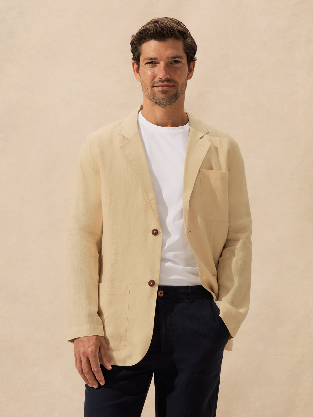 A man wearing a light beige linen blazer over a white t-shirt and dark navy trousers, standing against a textured beige background.