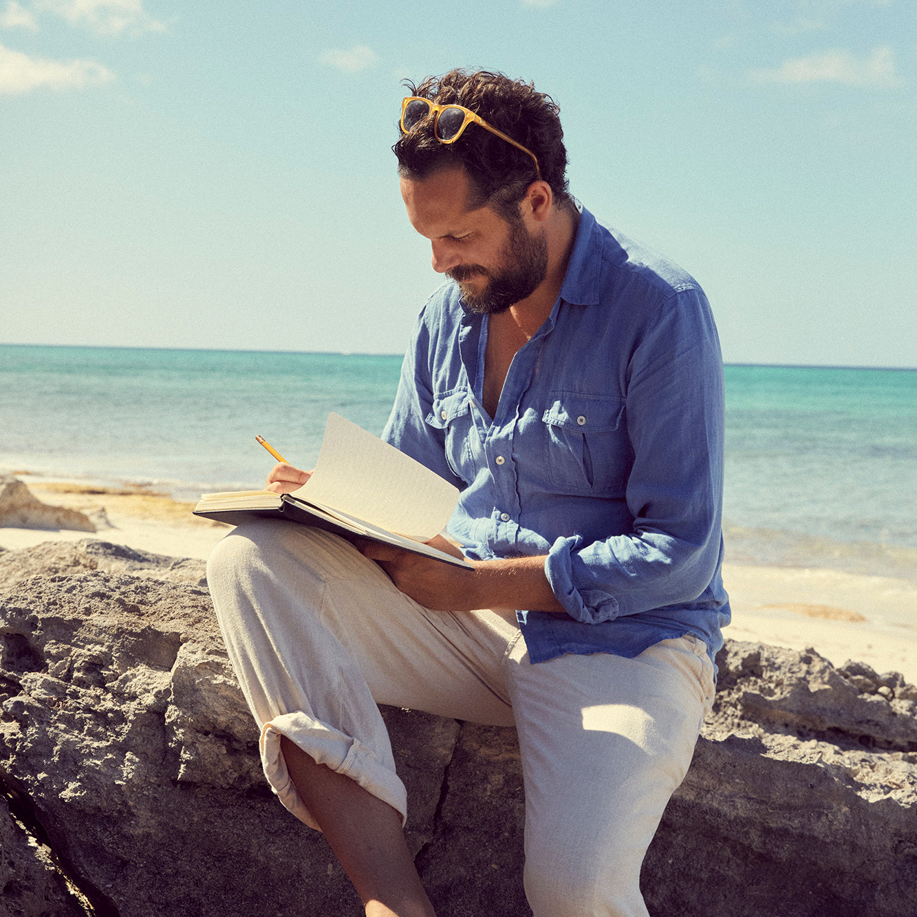 Oliver Tomalin with sunglasses on his head sits on a rock by the ocean, writing in a notebook. He is wearing a blue button-up shirt and light beige pants.